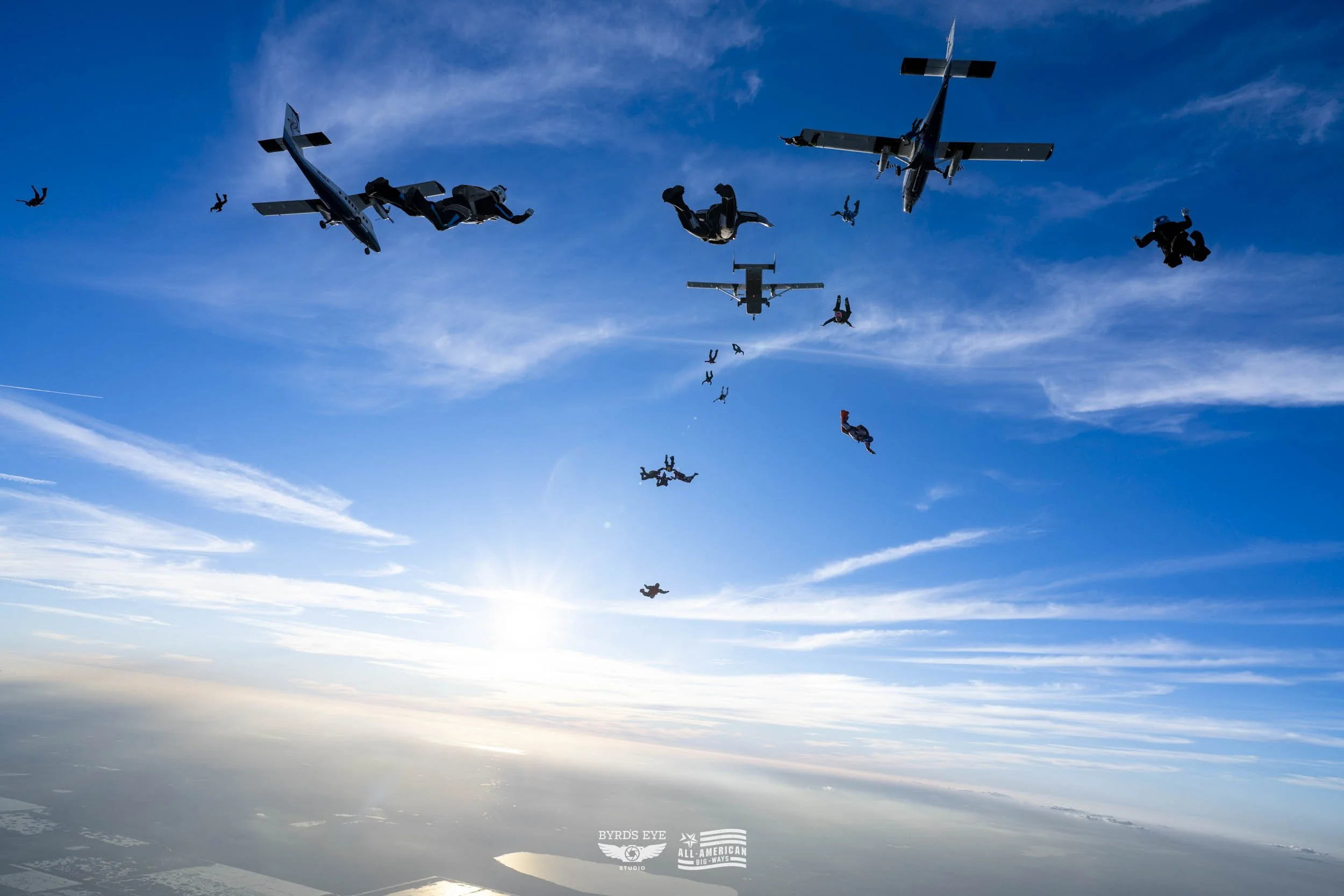 Skydivers and airplanes in a formation high above the clouds during daytime.