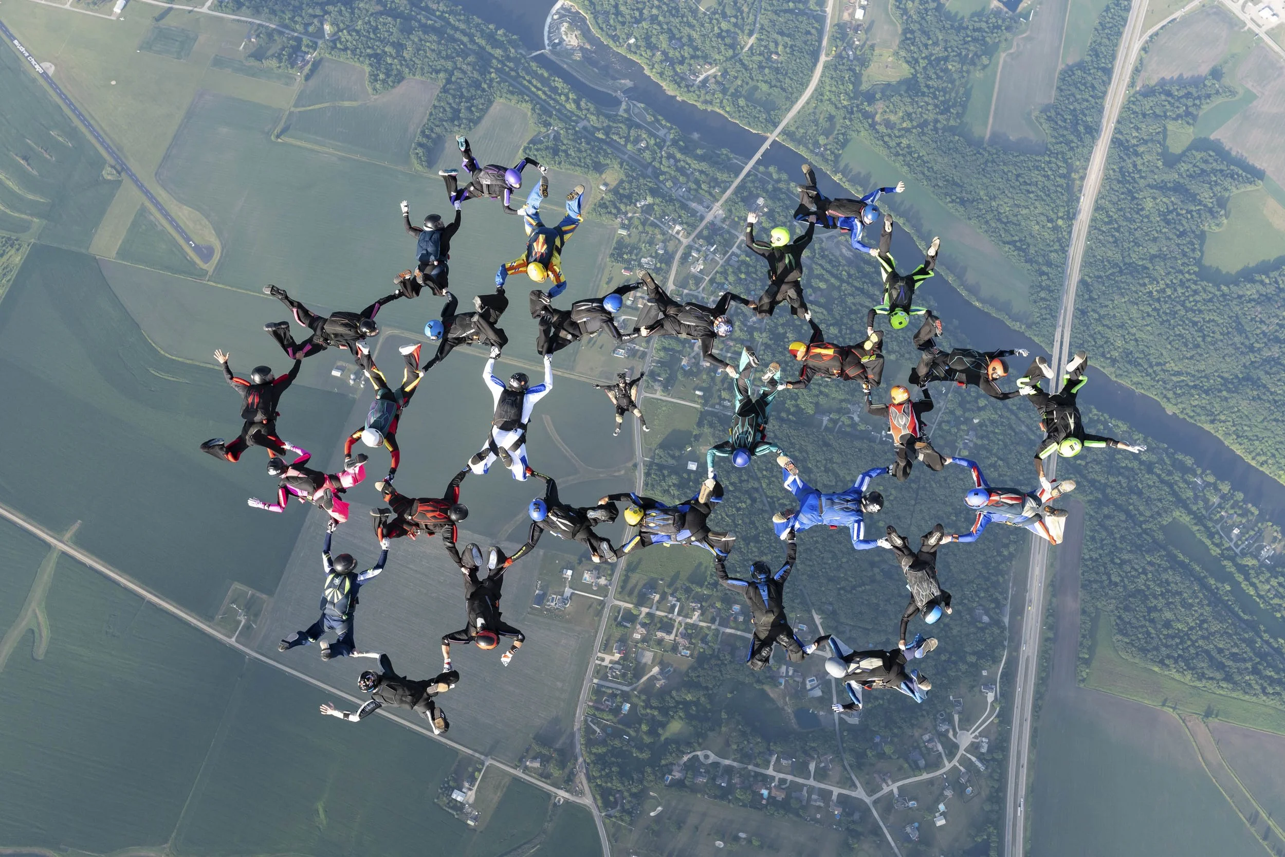 A large group of skydivers forming various shapes and patterns in the sky over a rural area with fields, roads, and a river below.