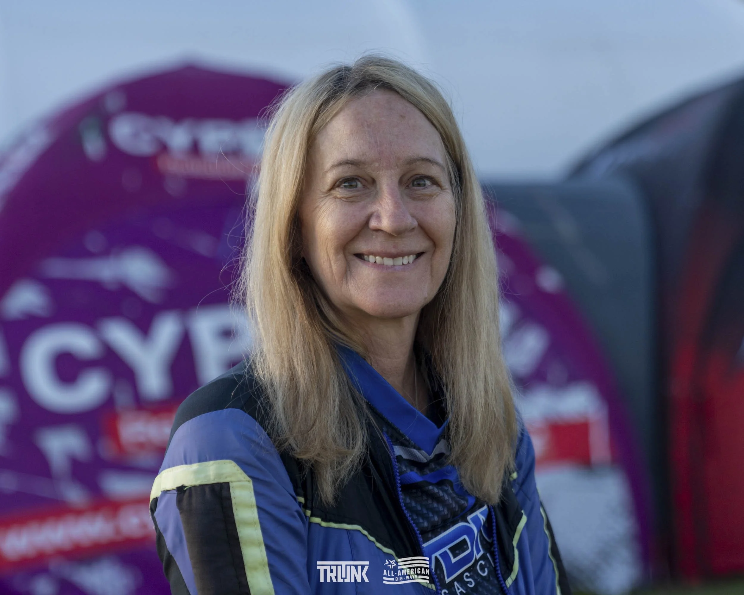 A smiling woman in a blue and black sports jacket with a logo, standing outdoors with blurred purple and black tents or structures in the background.