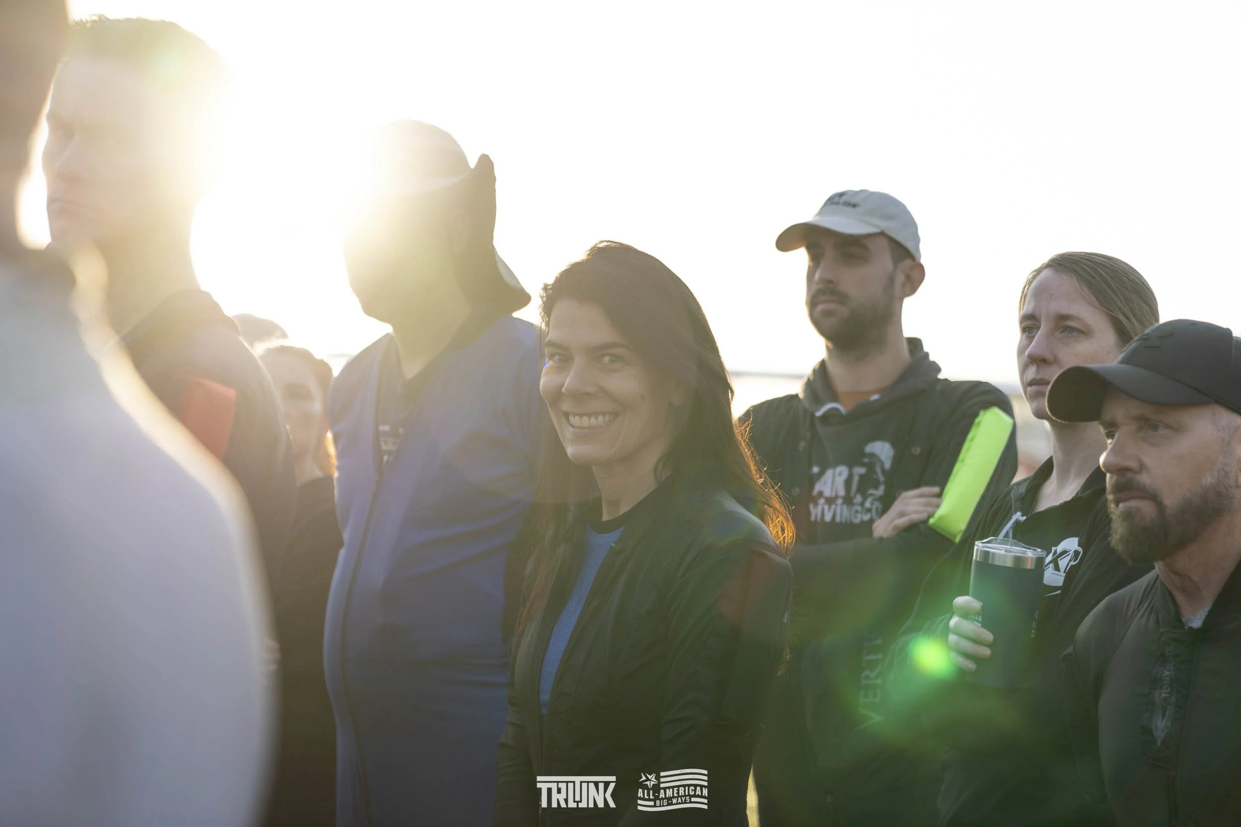 A group of people gathered outdoors with the sun setting behind them. A woman smiling at the camera is in the center, while others look in different directions, some holding drinks. The scene appears to be a casual or social event.