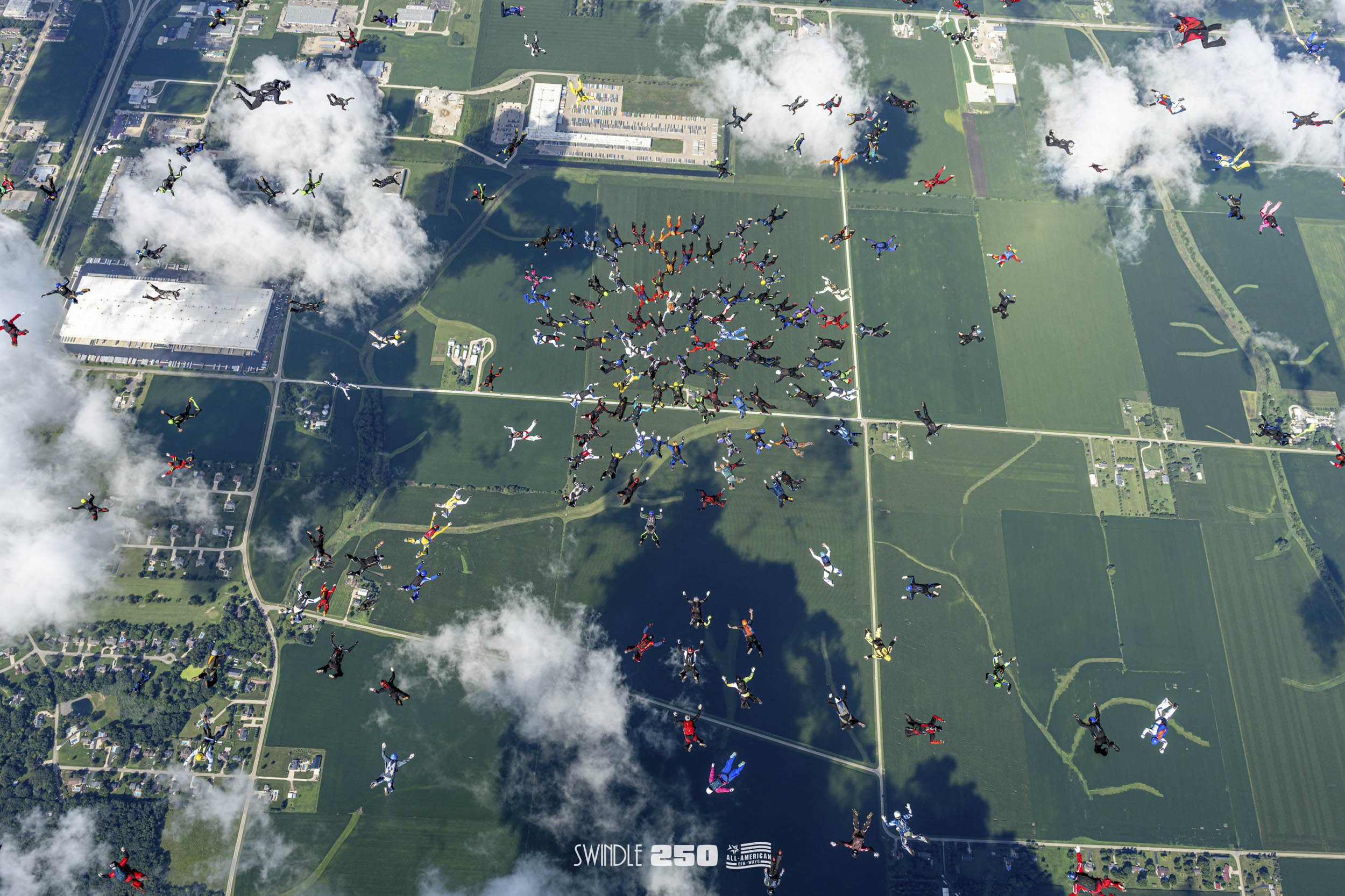 Hundreds of skydivers in colorful jumpsuits descending over a rural landscape with green fields, clouds, and a few buildings.
