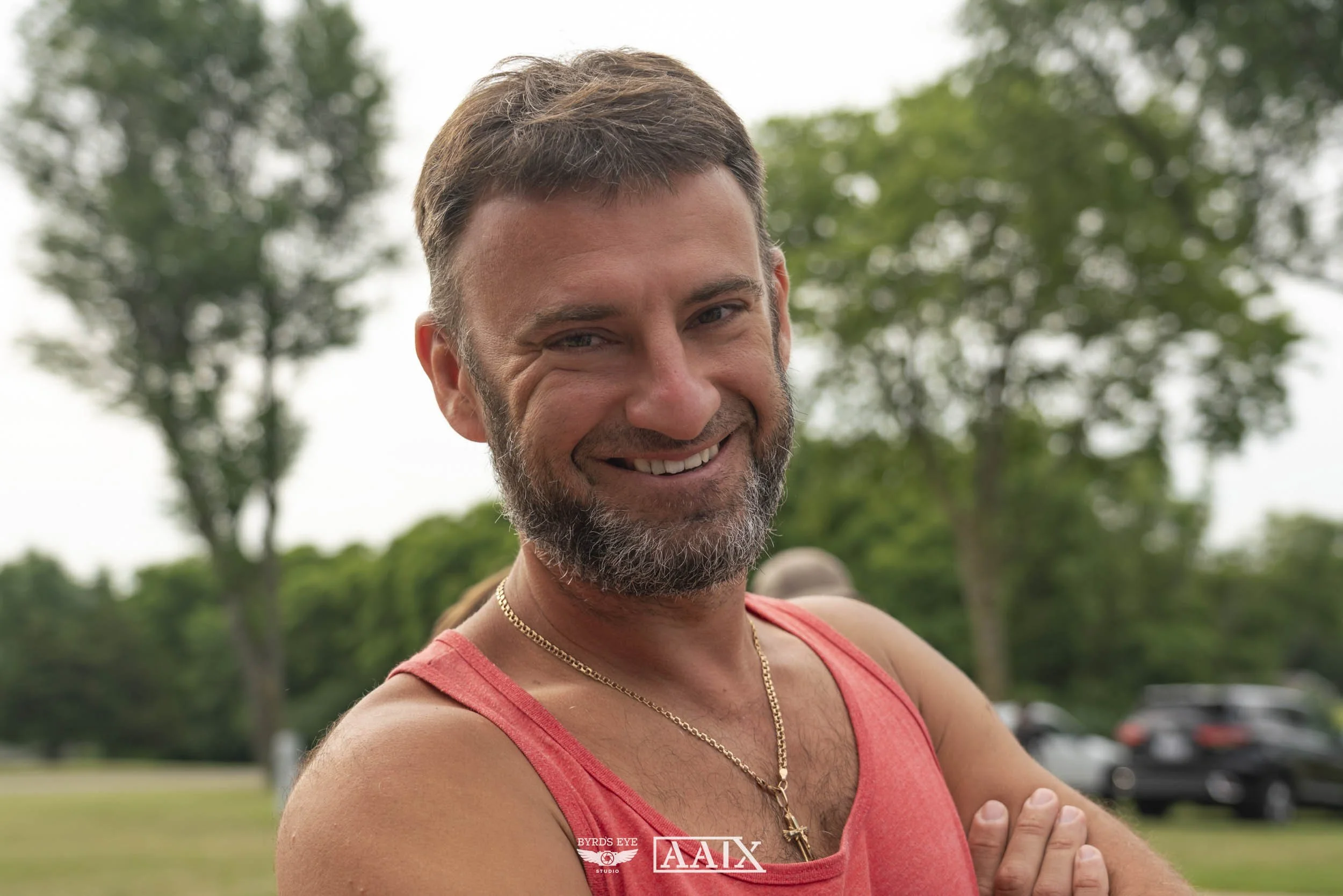 A smiling man with a beard and short brown hair, wearing a red tank top and gold chain necklace, standing outdoors with trees and cars in the background.