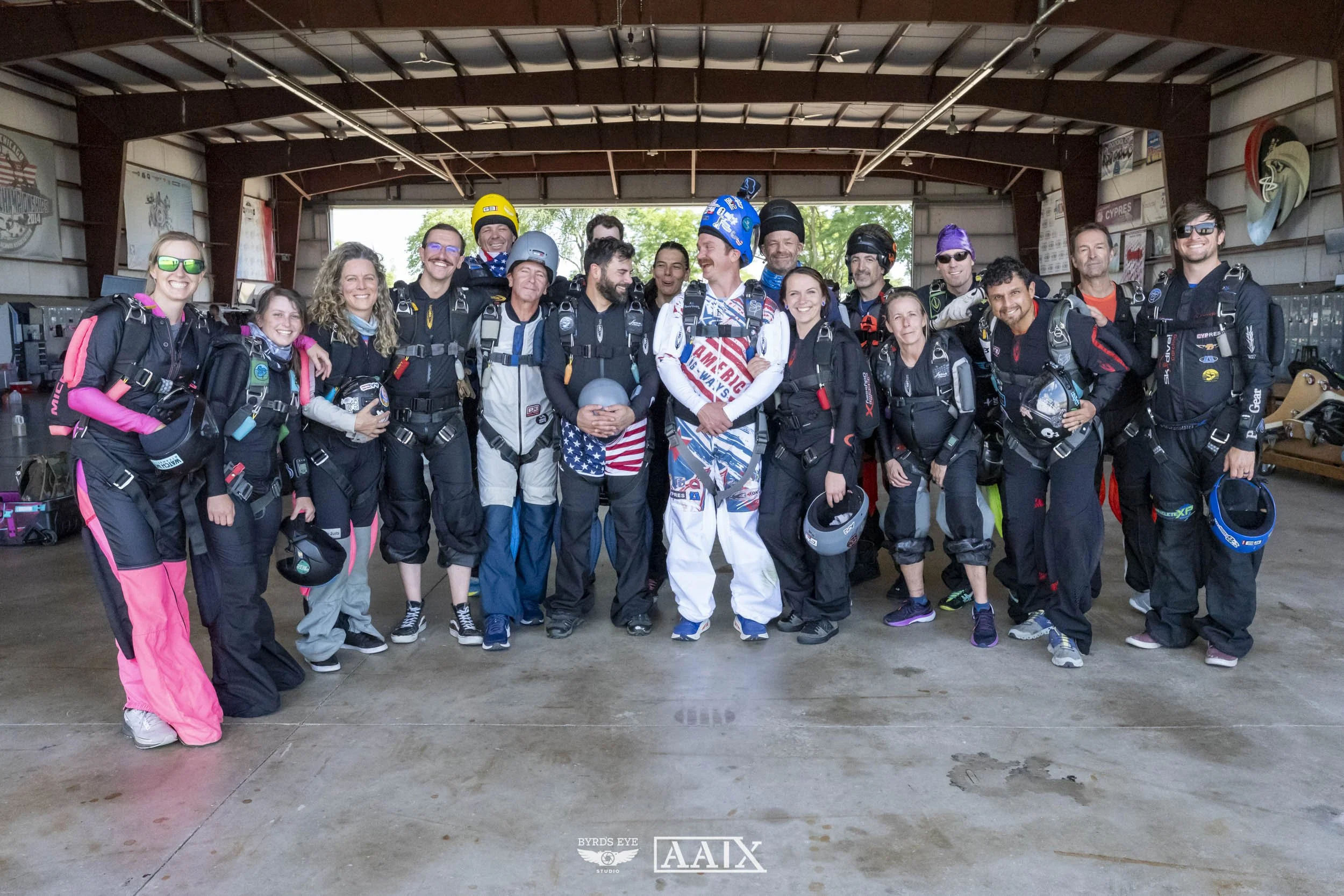 A group of skydivers dressed in jumpsuits and holding helmets, standing together in a hangar with large smiling faces, celebrating their skydiving experience.