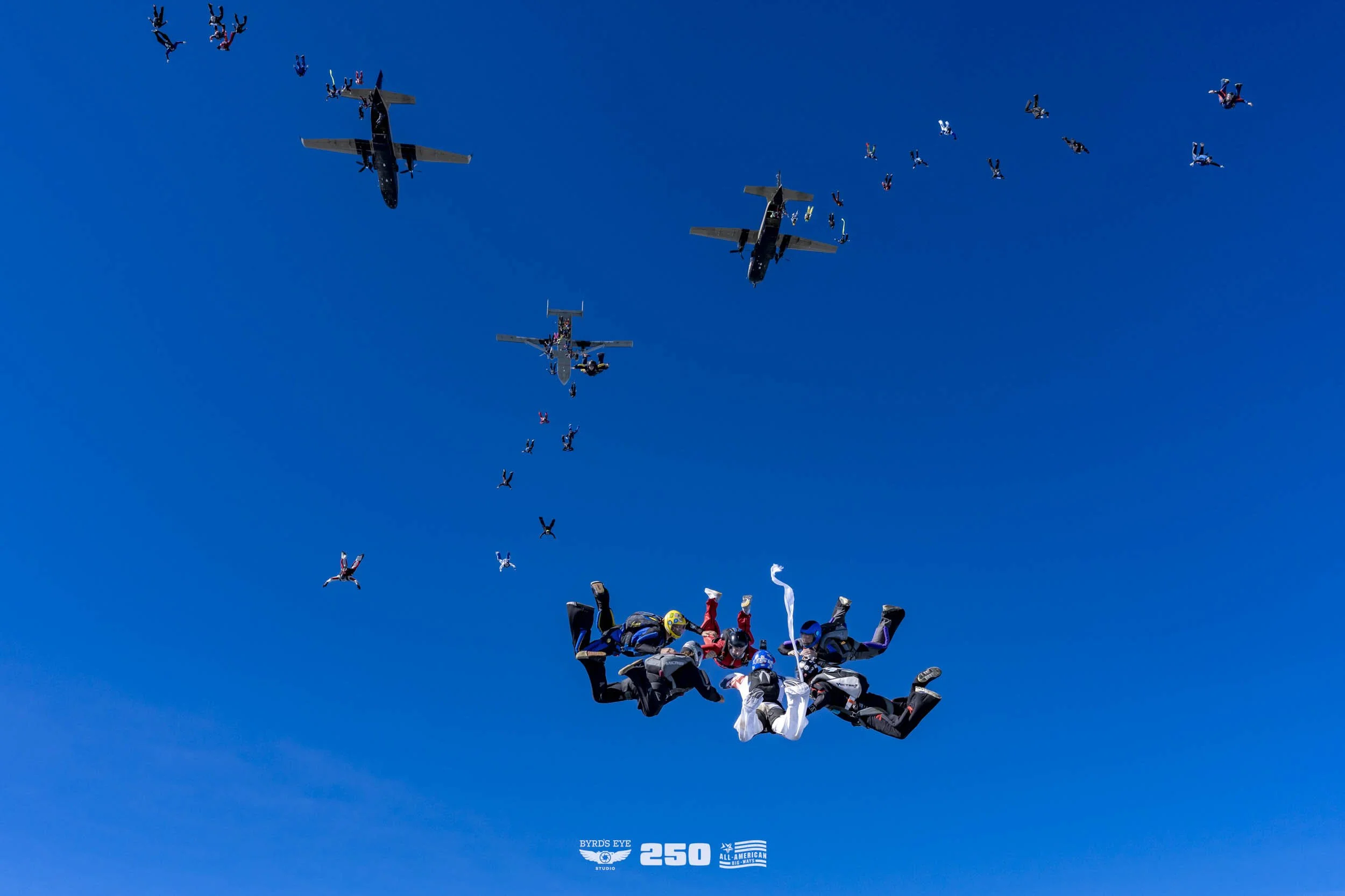 Skydivers in freefall formation below airplanes flying in a clear blue sky.