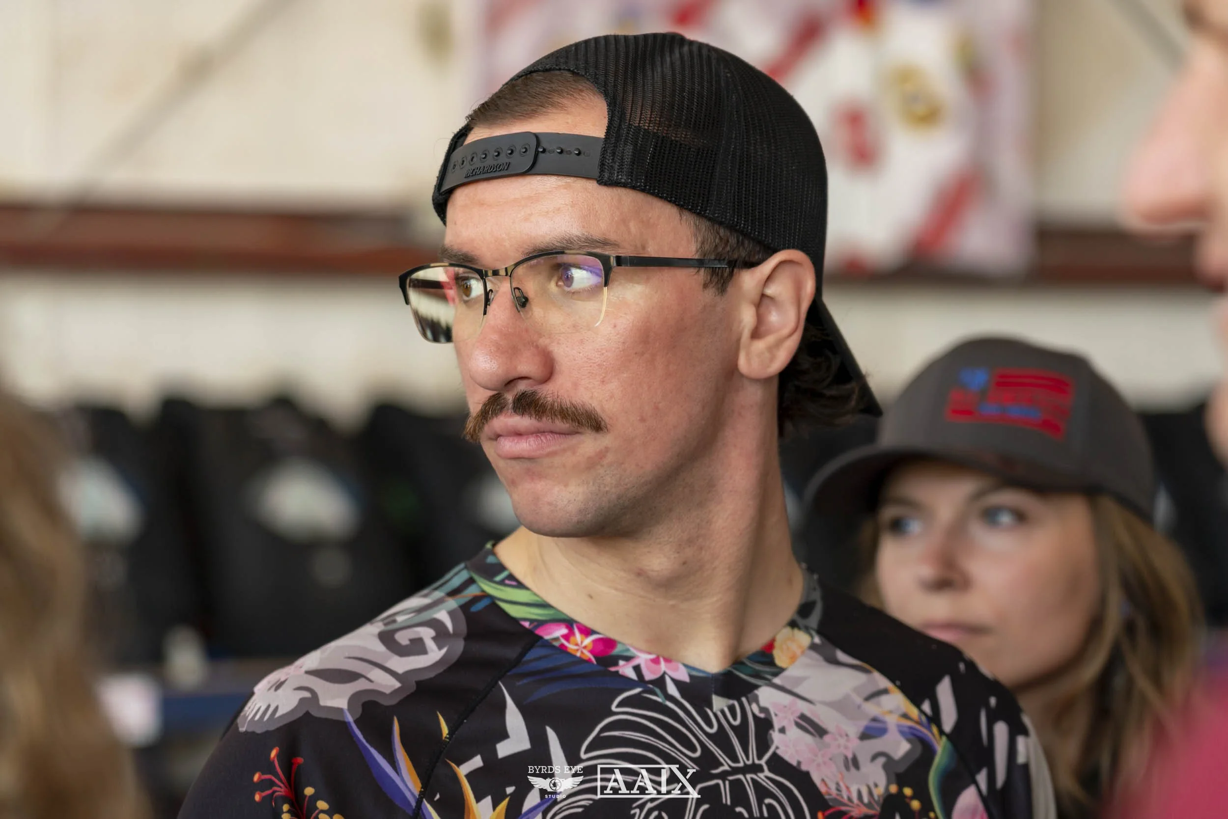 A young man with glasses and a mustache, wearing a black baseball cap backwards and a colorful shirt, looking to the side. There are two women in the background, one wearing a gray cap and the other with long hair.