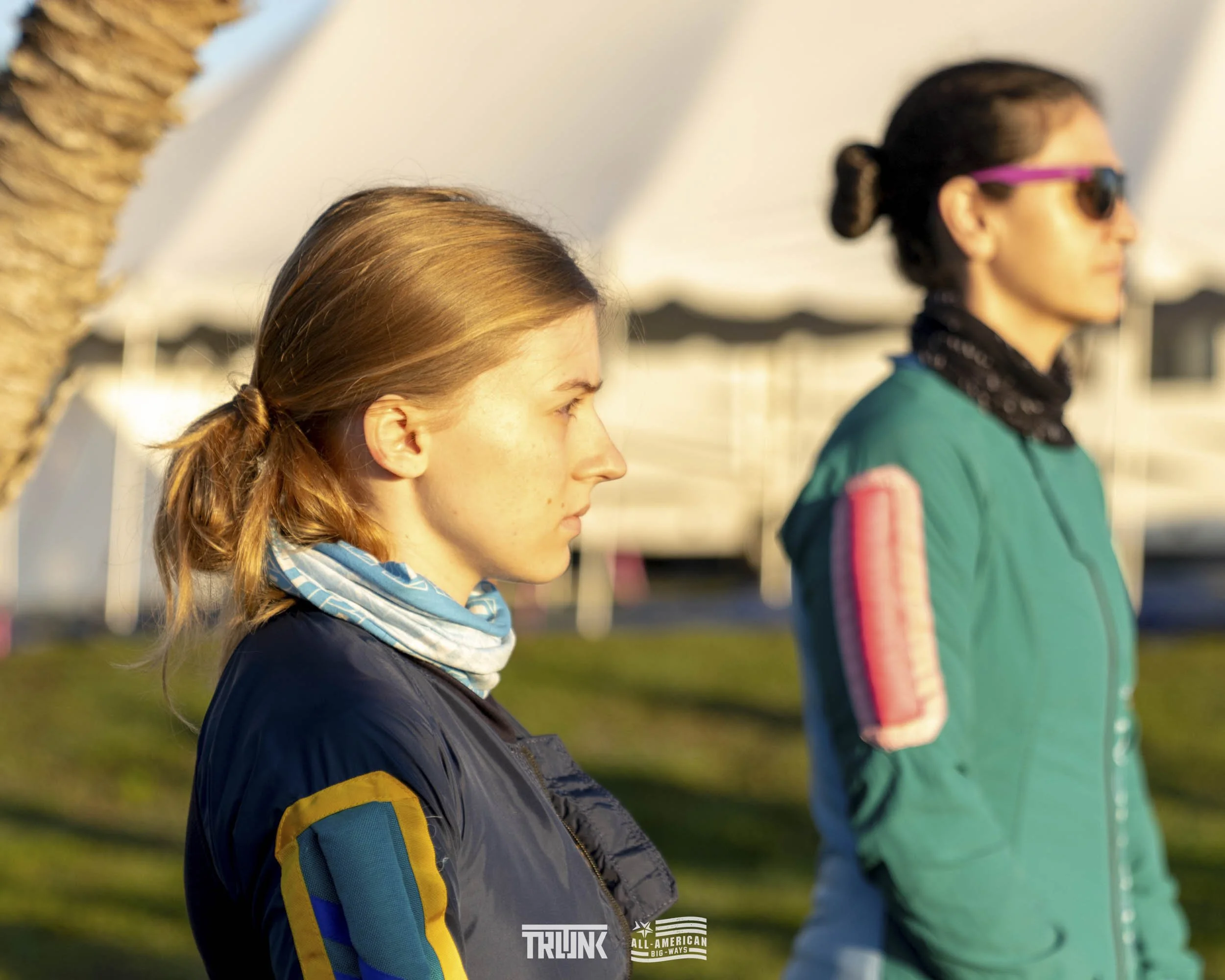 Two women standing outdoors, side by side, during golden hour. The woman in the foreground has light brown hair tied in a loose ponytail, and is wearing a black jacket with colored patches on the arm and a light-colored neck gaiter. The woman in the 