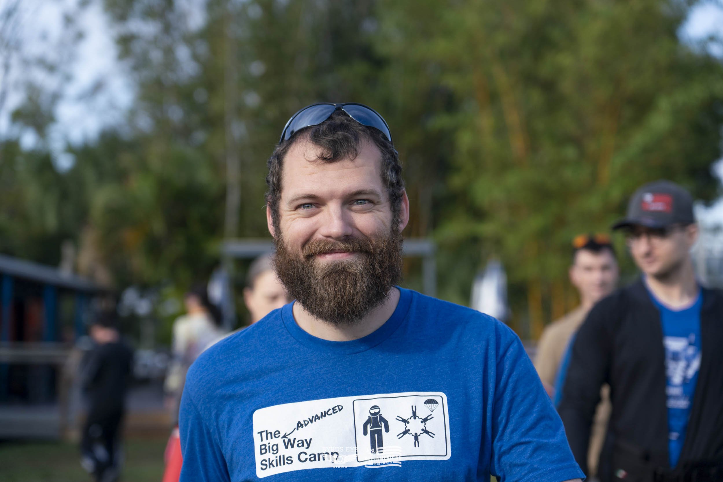 A man with a beard wearing sunglasses on his head and a blue t-shirt with a logo and text, smiling outdoors with other people in the background.