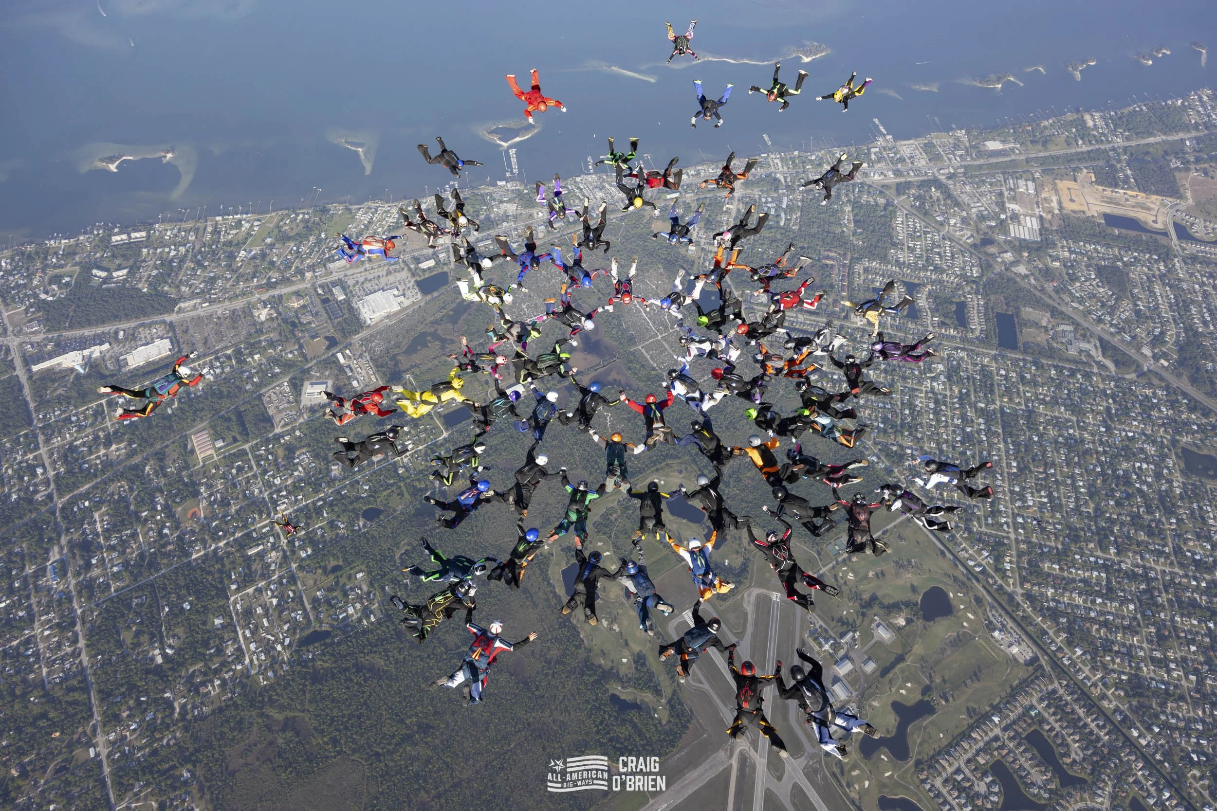 Skydivers in colorful jumpsuits forming a massive star shape as they fall over a city landscape with water bodies.