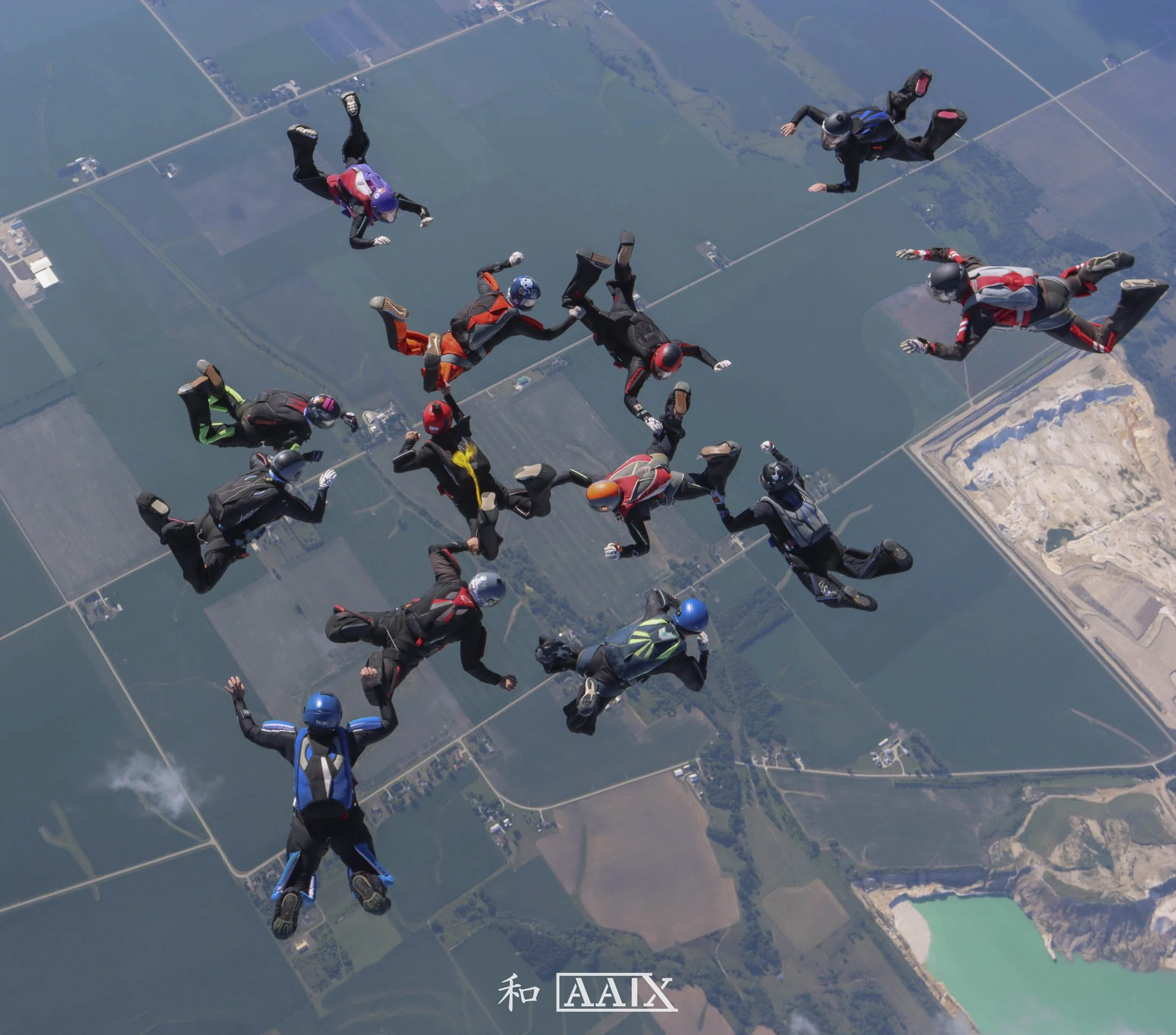 A group of skydivers in free fall over a patchwork of fields and water bodies, holding hands in a circle.