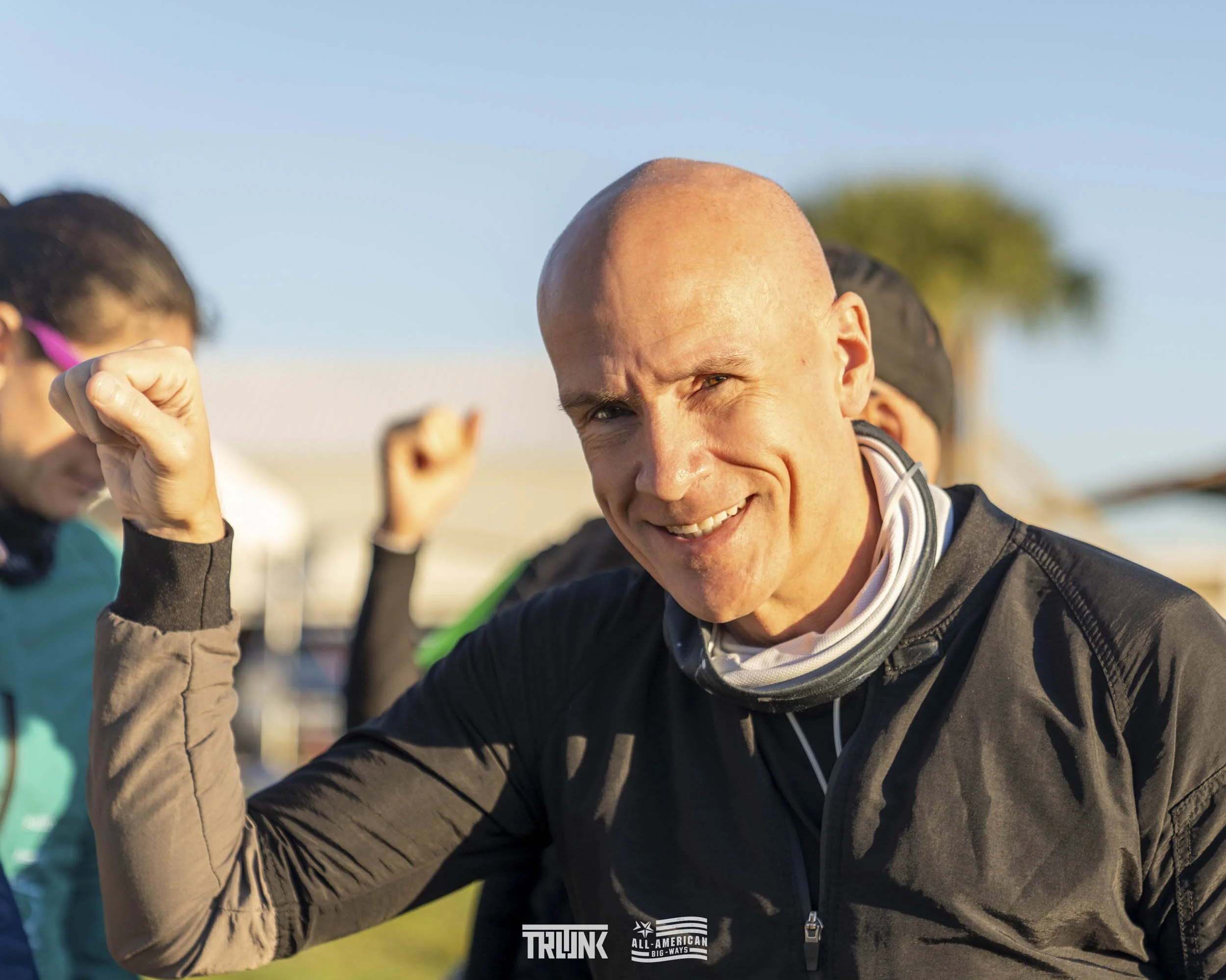 A smiling man with a bald head, wearing a black jacket and a neck gaiter, flexing his bicep at an outdoor event with others in the background, some raising their fists.