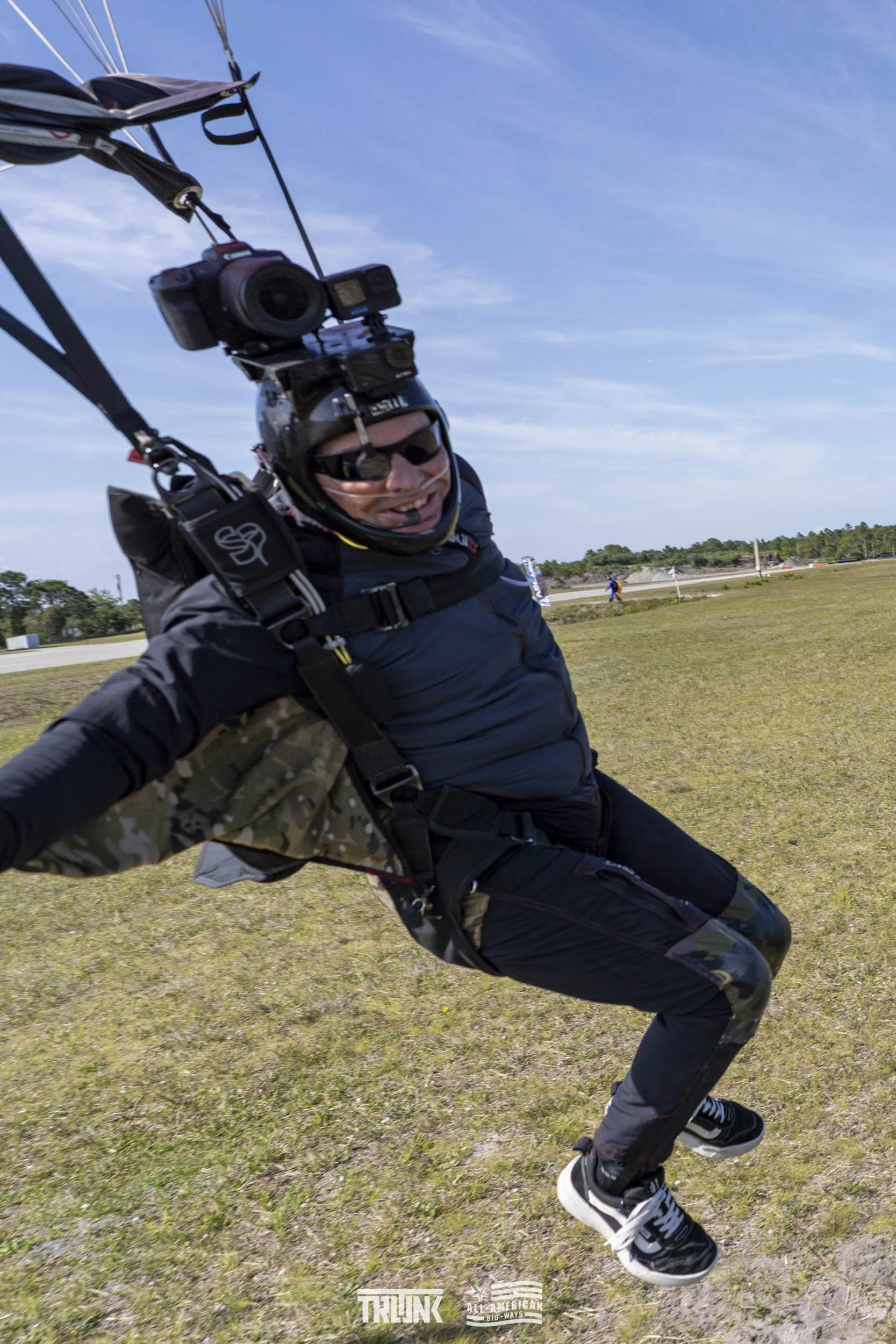 A man in a harness and helmet with a camera, mid-air during a parachuting jump, smiling and wearing sunglasses, with an open field and sky in the background.