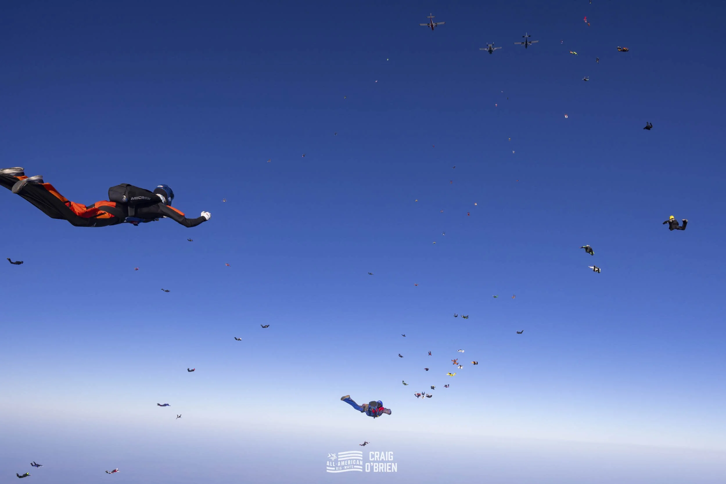 Multiple skydivers descending from the sky with a few planes overhead against a blue sky.