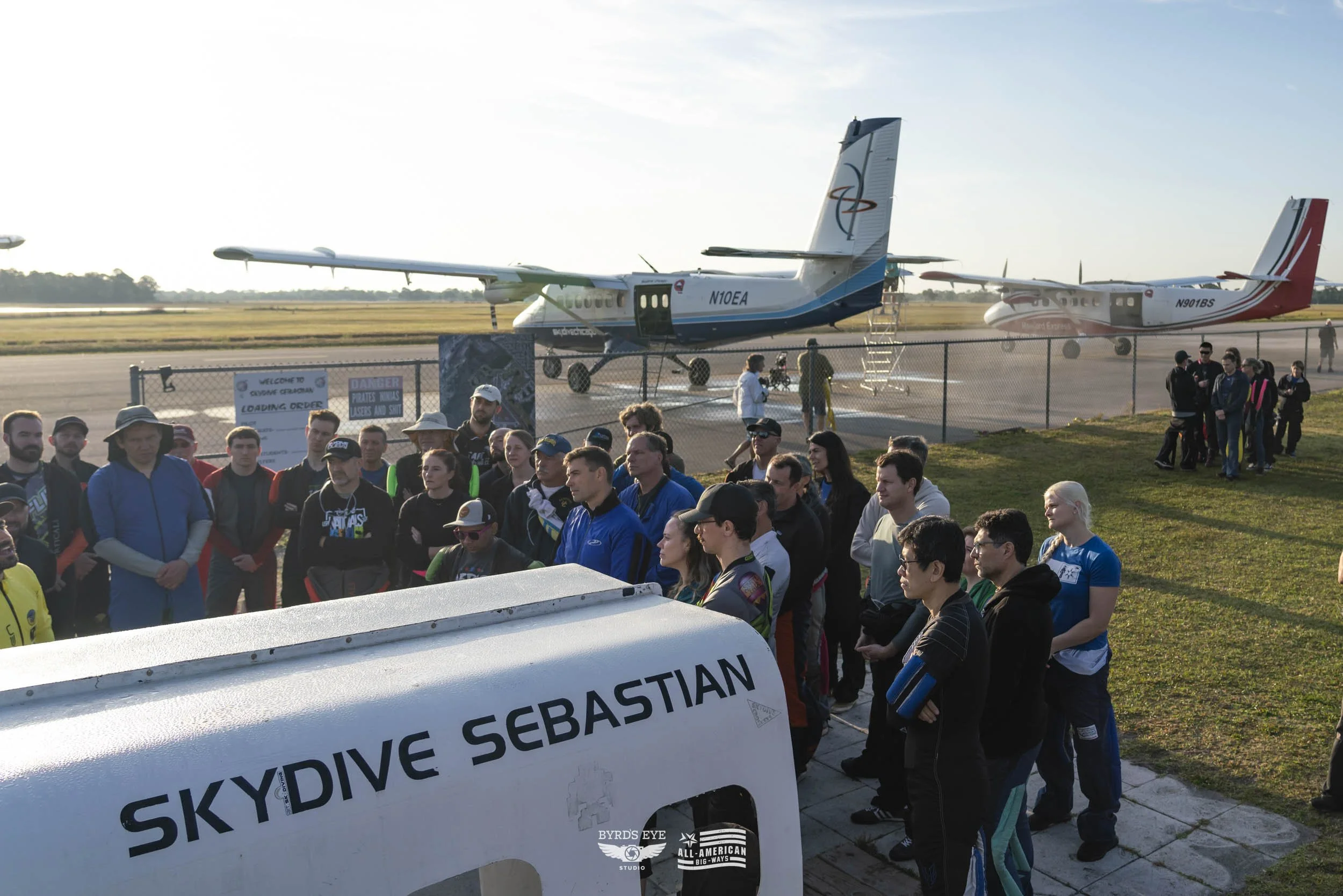 A group of people gathered at an airport with airplanes in the background, some standing near a fence and a sign that reads 'SKYDIVE SEBASTIAN.' The scene appears to be part of a skydiving event or tour.