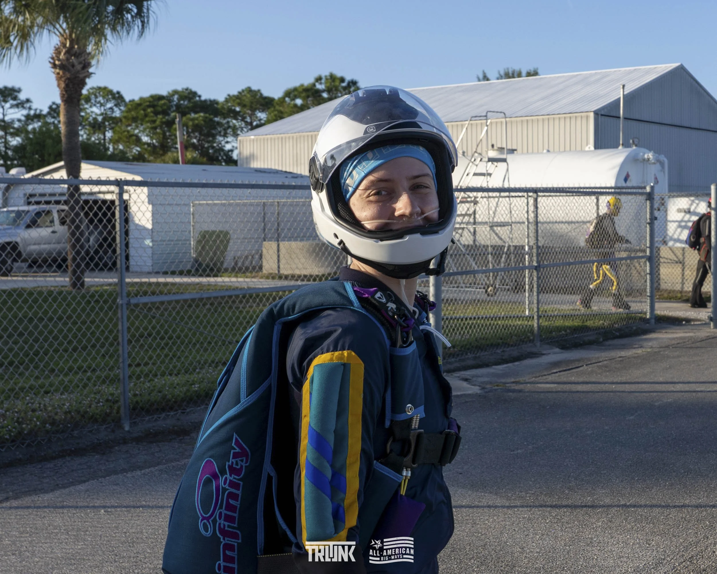 Person wearing a motorcycle helmet and a blue jacket, carrying a backpack with a colorful patch, standing outdoors near a chain-link fence, with a large white building, trees, and people in the background.
