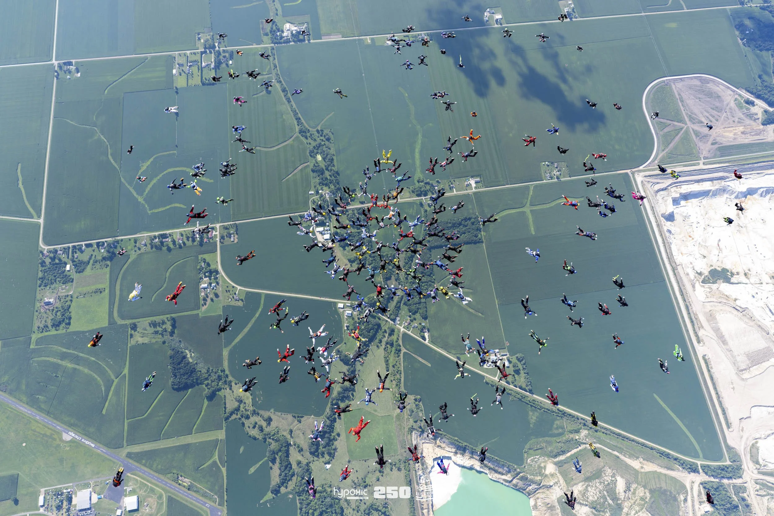 Skydivers jumping out of an airplane over a green landscape with fields, roads, and a large water body, forming a heart shape in mid-air.