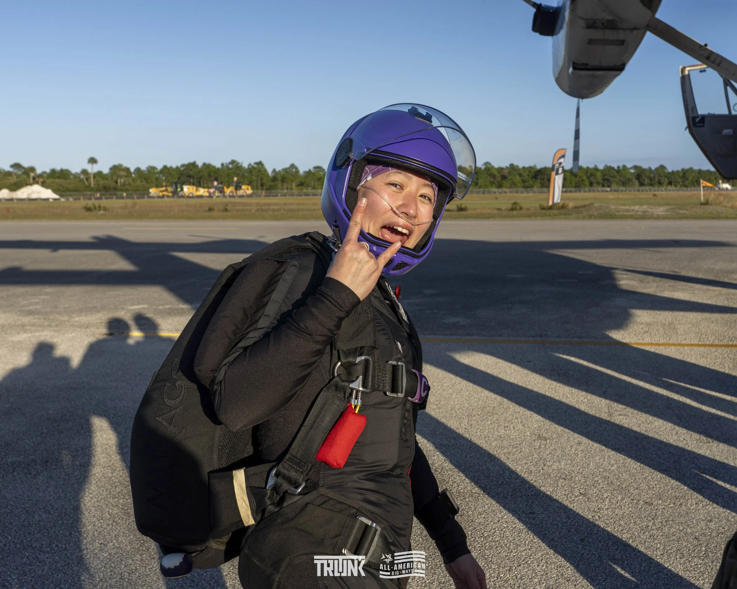 A woman in a purple helmet and black jumpsuit making a rock sign with her hand at an airport tarmac, with a large aircraft and luggage tractors in the background.