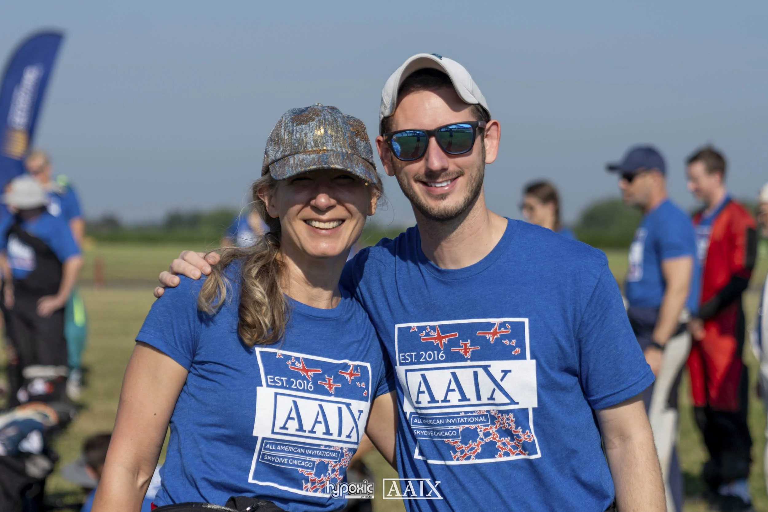 Two smiling people wearing blue shirts with aviation-themed designs, standing outdoors at an event with other people in the background.