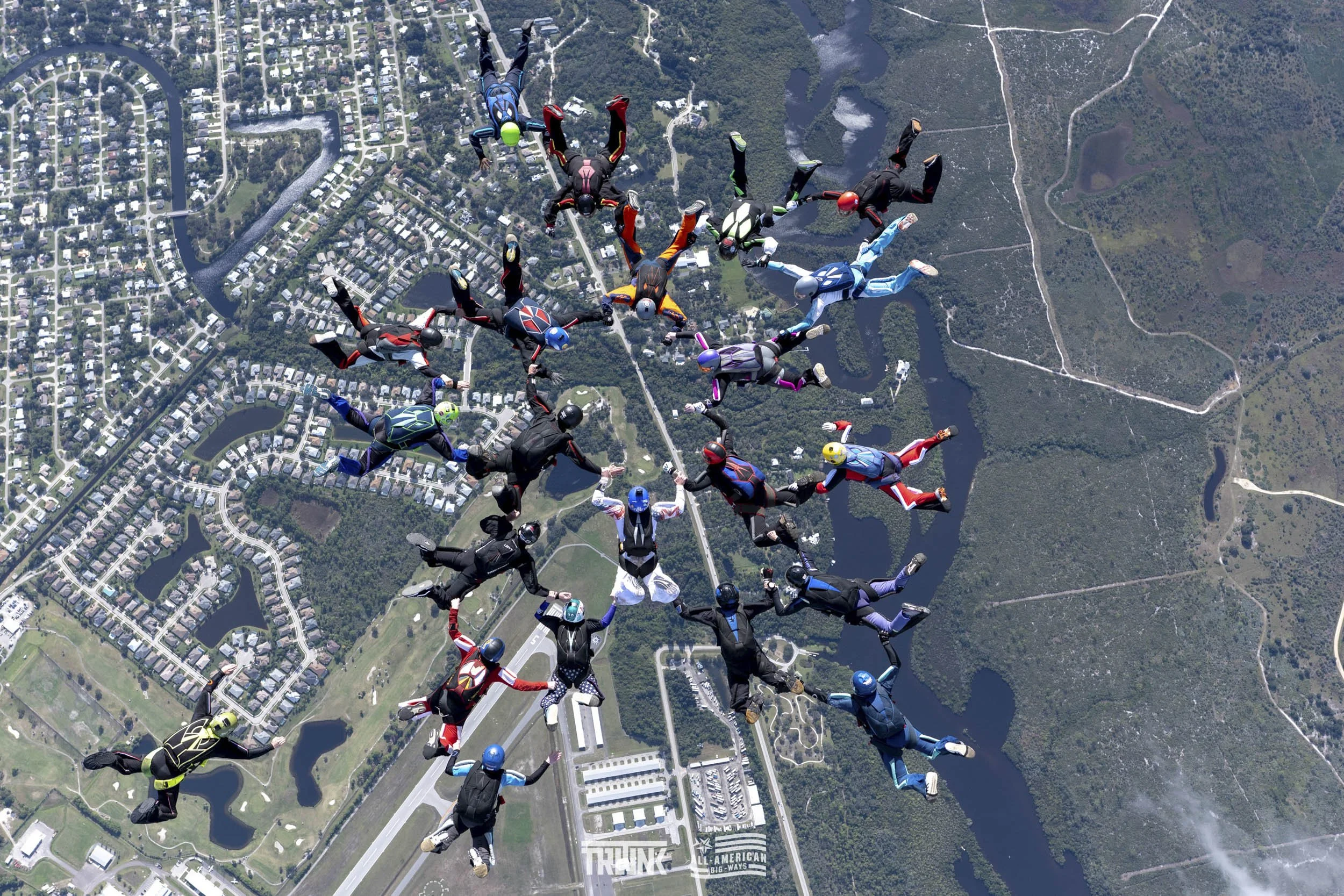 Group of skydivers in colorful jumpsuits and helmets forming a star shape during free fall over an urban area with lakes, roads, and green parks.