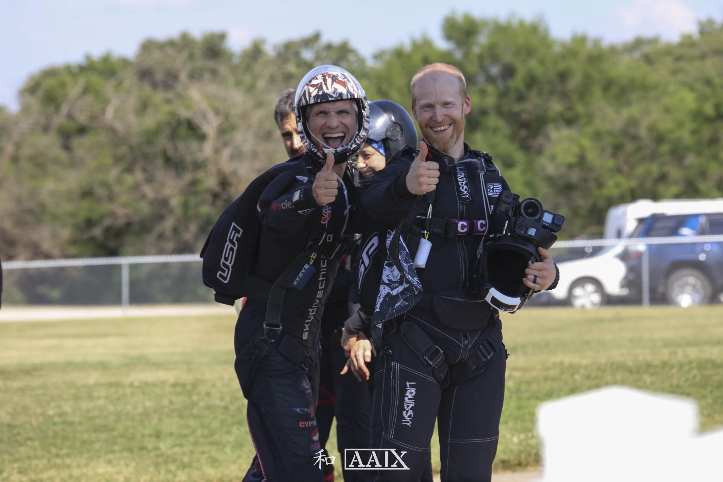 Four skydivers in jumpsuits and helmets standing on grass, smiling and giving thumbs up after a jump, with trees and vehicles in the background.