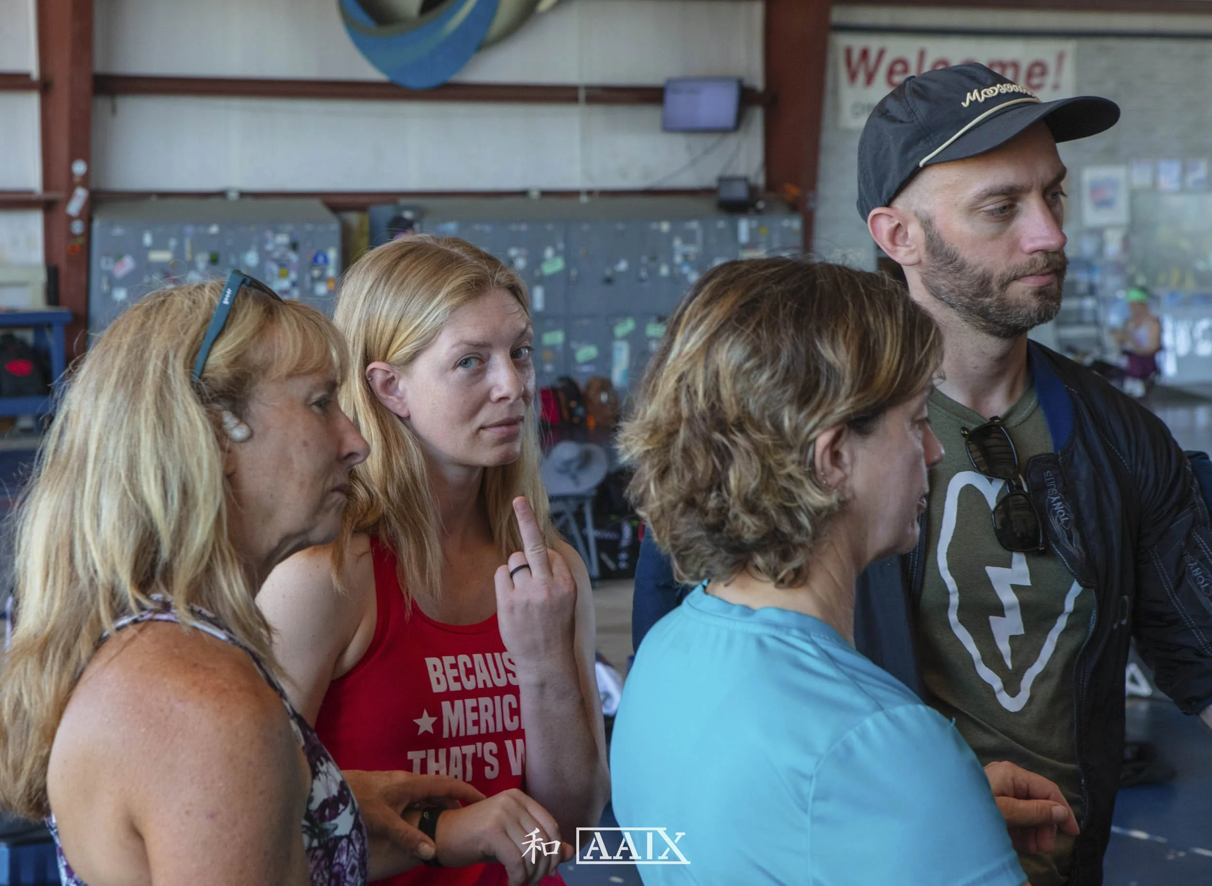 A group of four adults, three women and one man, are gathered closely together in an indoor setting with a background of lockers and a welcome sign. They appear to be listening attentively or waiting.