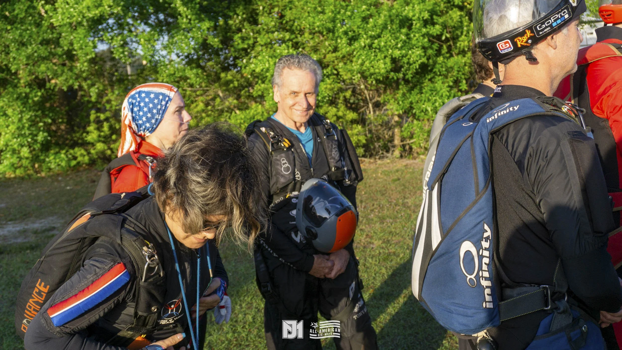 Group of skydivers in black jumpsuits with backpacks, some with helmets, preparing for a jump outdoors with a green backdrop.