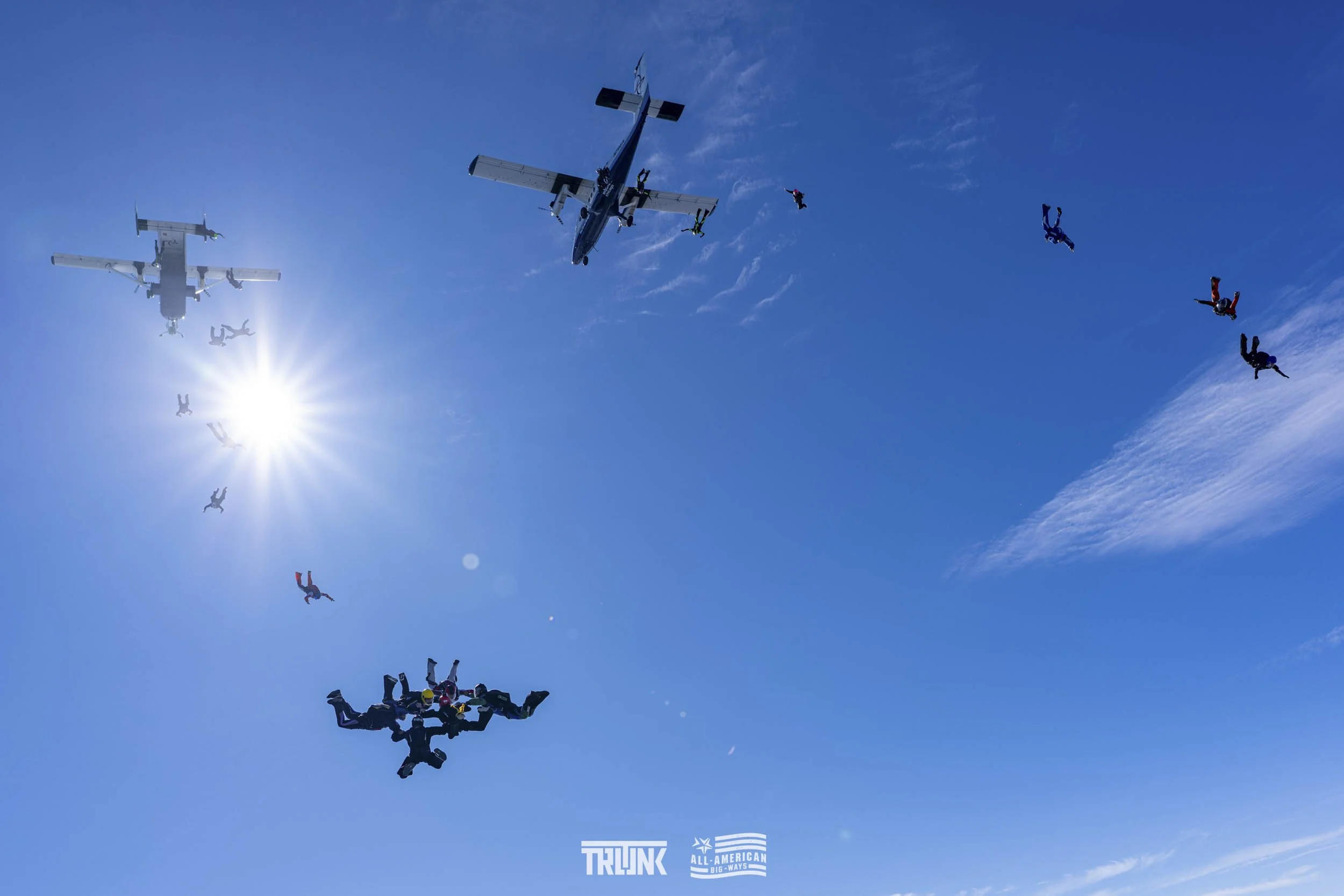 Skydivers in free fall formations with two airplanes flying overhead on a bright, sunny day.