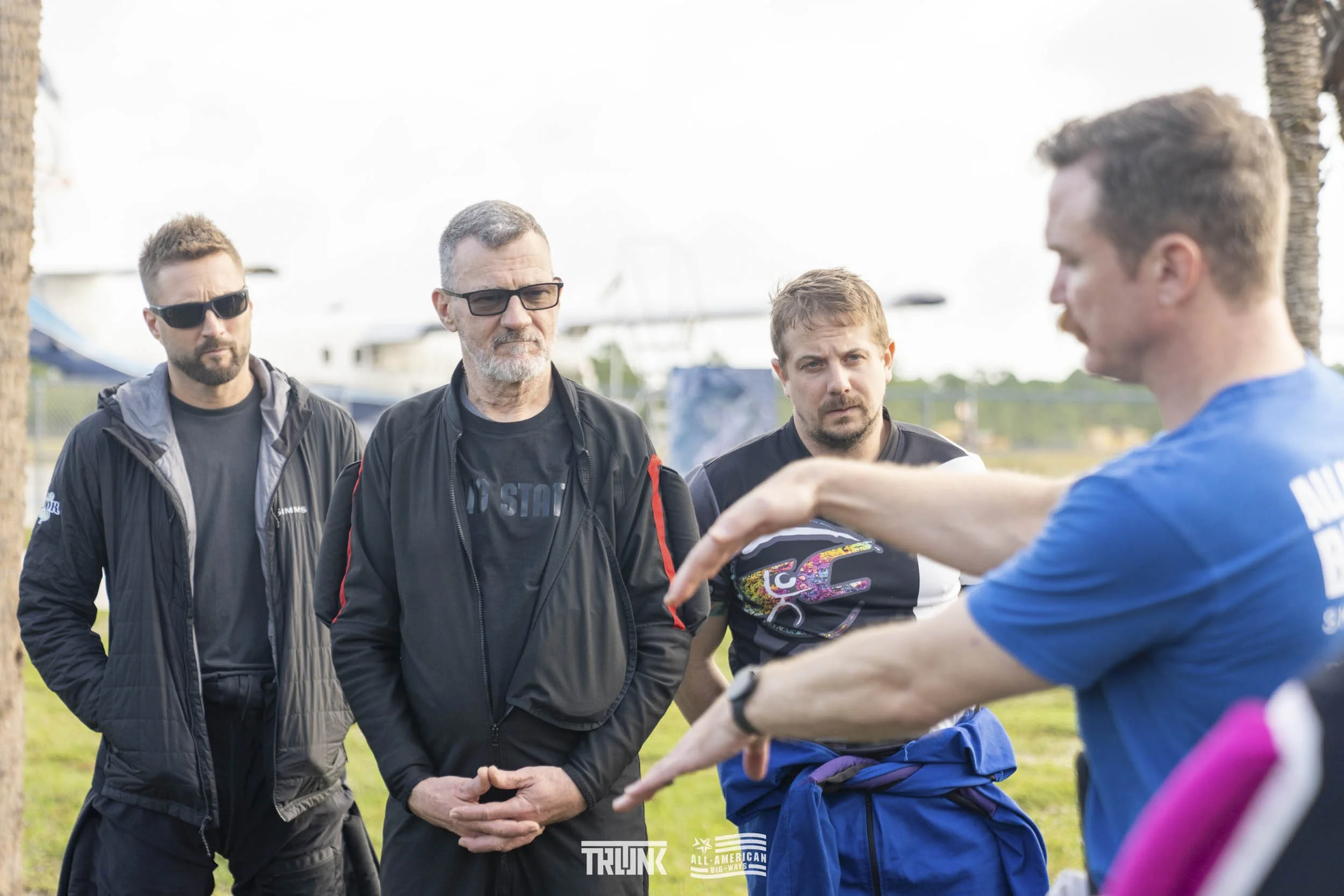 Four men standing outdoors, listening to a man in a blue shirt who is speaking and gesturing with his hands. Three of the men are wearing black jackets, one has a gray jacket, and one has a black T-shirt. Two men are wearing sunglasses. Background sh