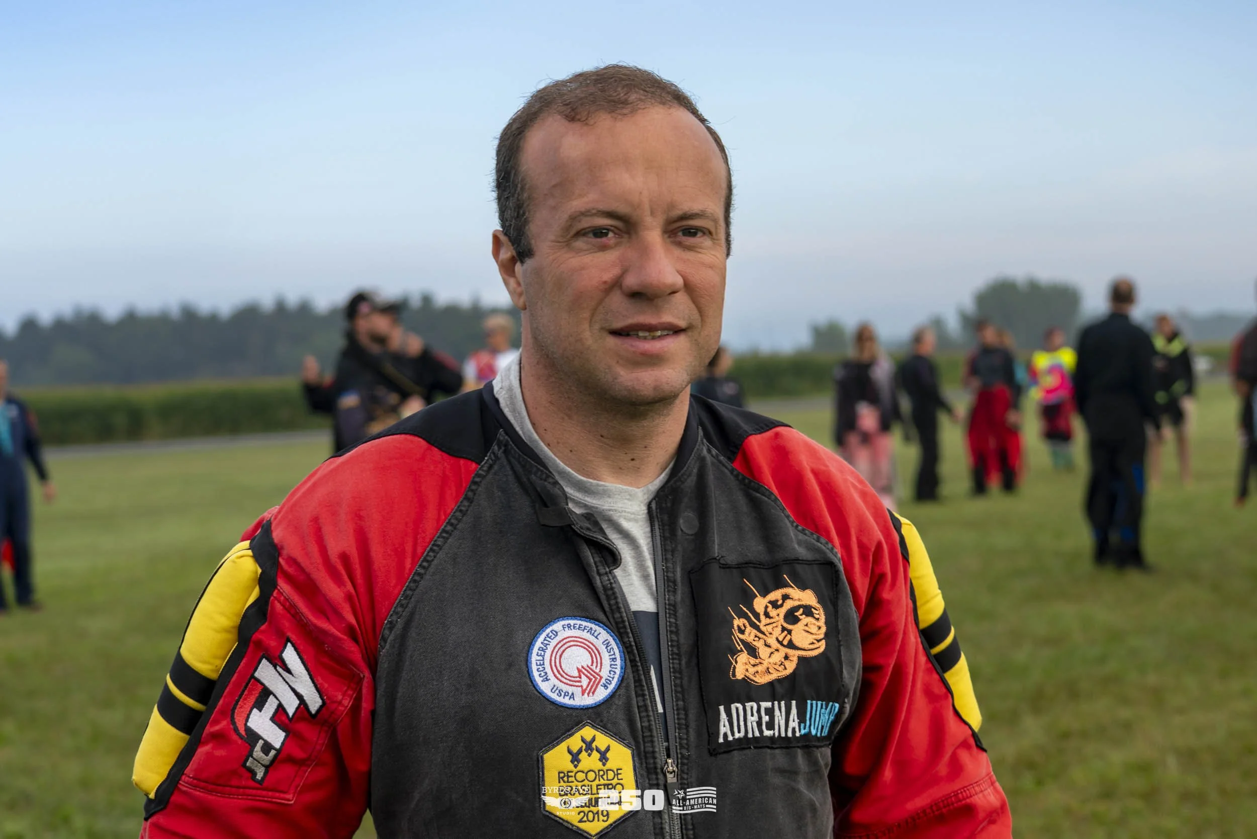 A man in a racing jumpsuit standing outdoors at a skydiving event, with other people in the background.