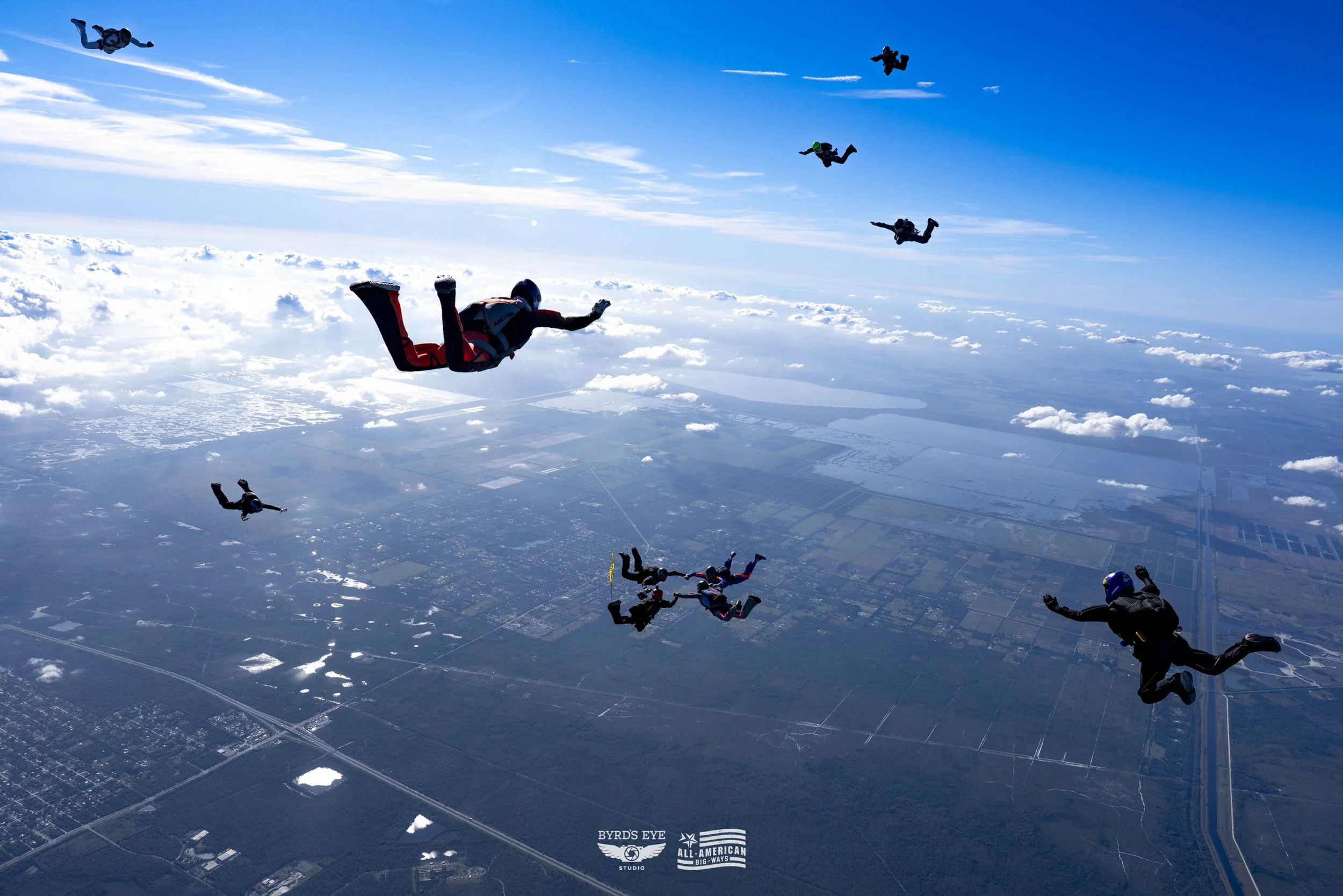 Skydivers free-falling through the sky above a landscape with clouds and fields below.