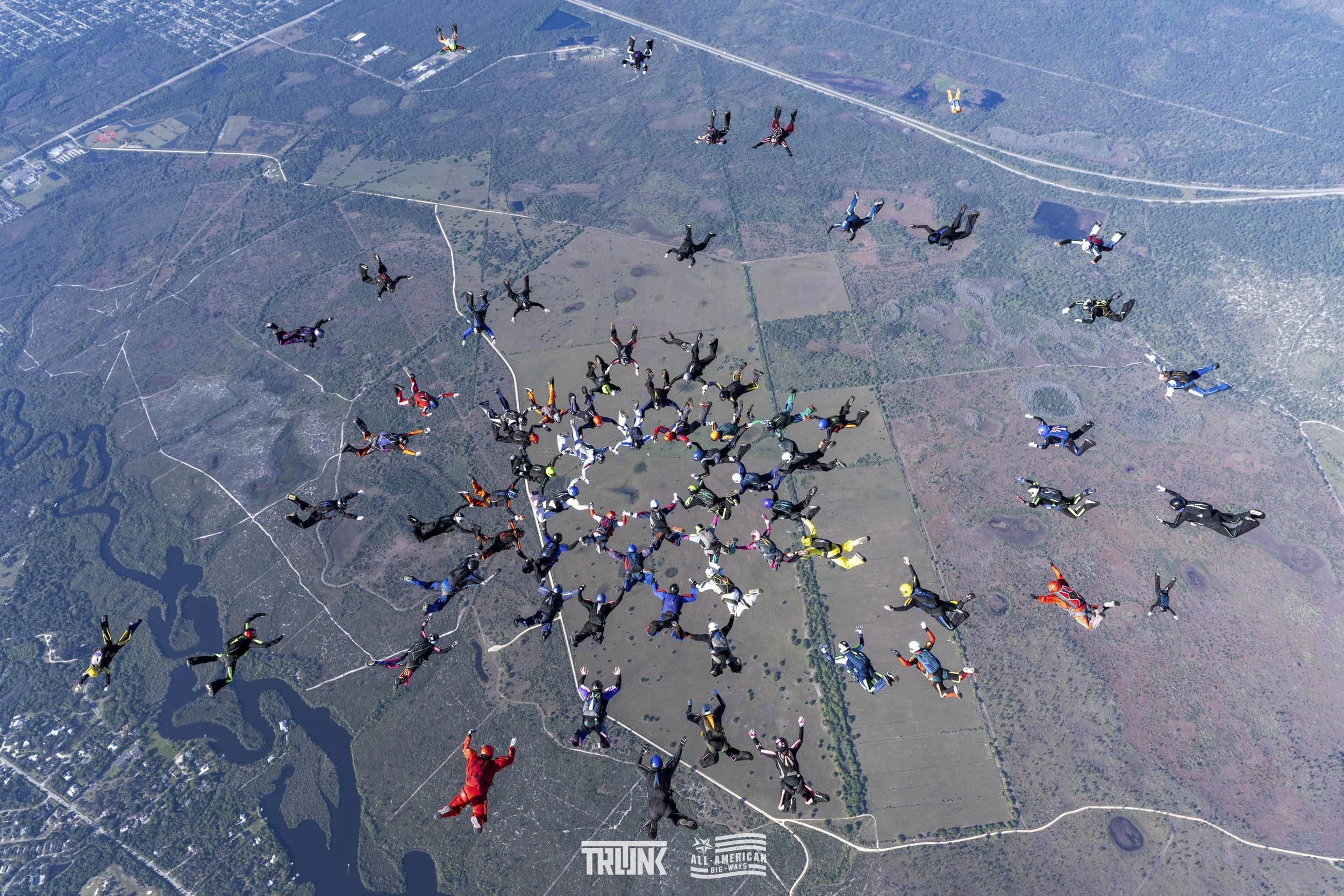 A large group of skydivers in colorful jumpsuits forming a circular formation while free-falling over a landscape with rivers, forests, and fields.