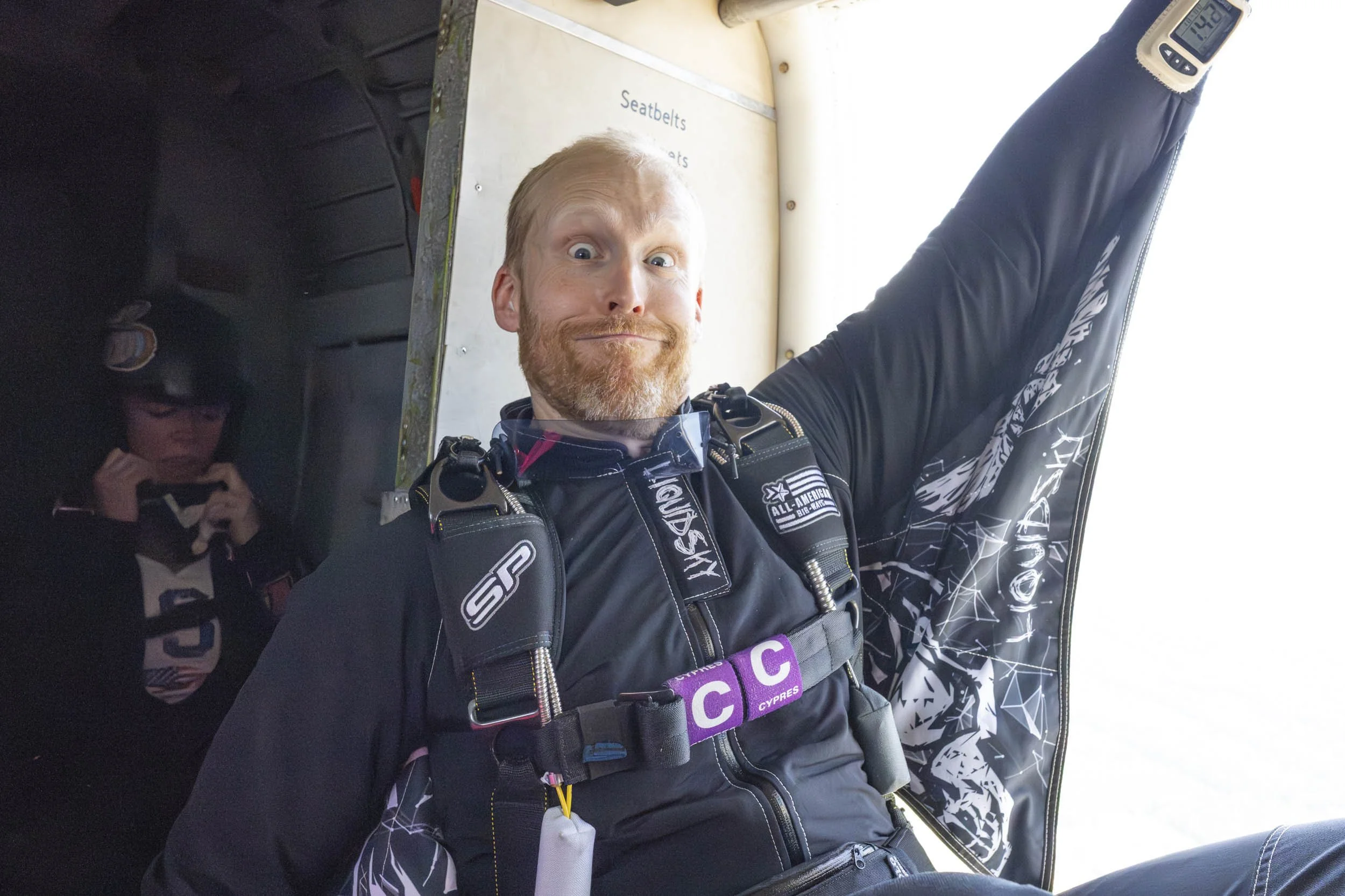 A man with a beard inside an airplane, wearing skydiving gear, smiling and raising an arm, with a woman in the background looking at her phone.