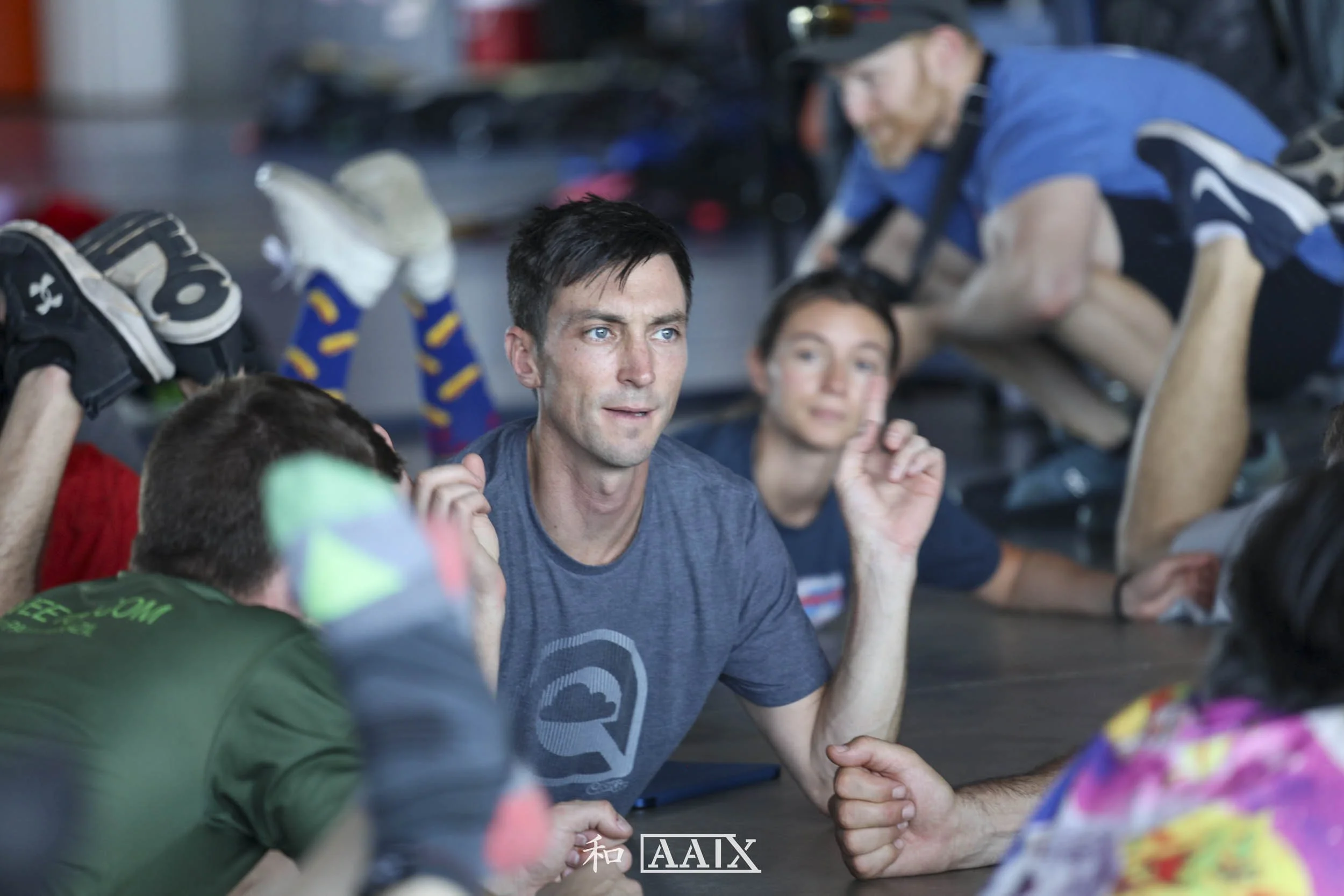 A group of people participating in a friendly arm wrestling match on the floor indoors, with some individuals sitting, lying down, or kneeling around them.