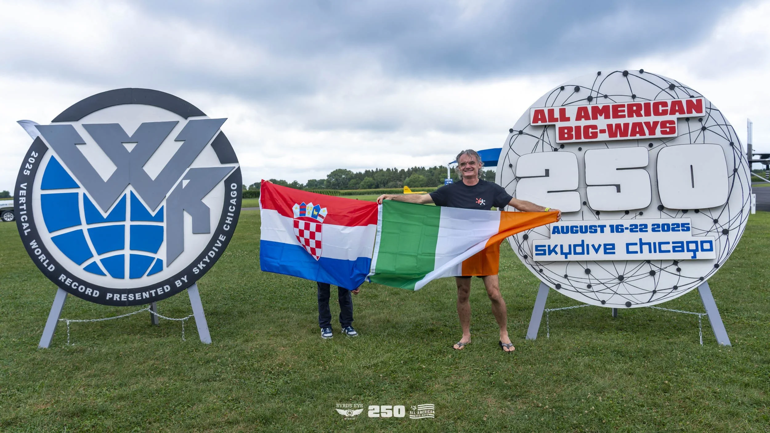 Person holding Croatian and Irish flags standing in front of signs for the 2025 World Record for skydiving in Chicago.