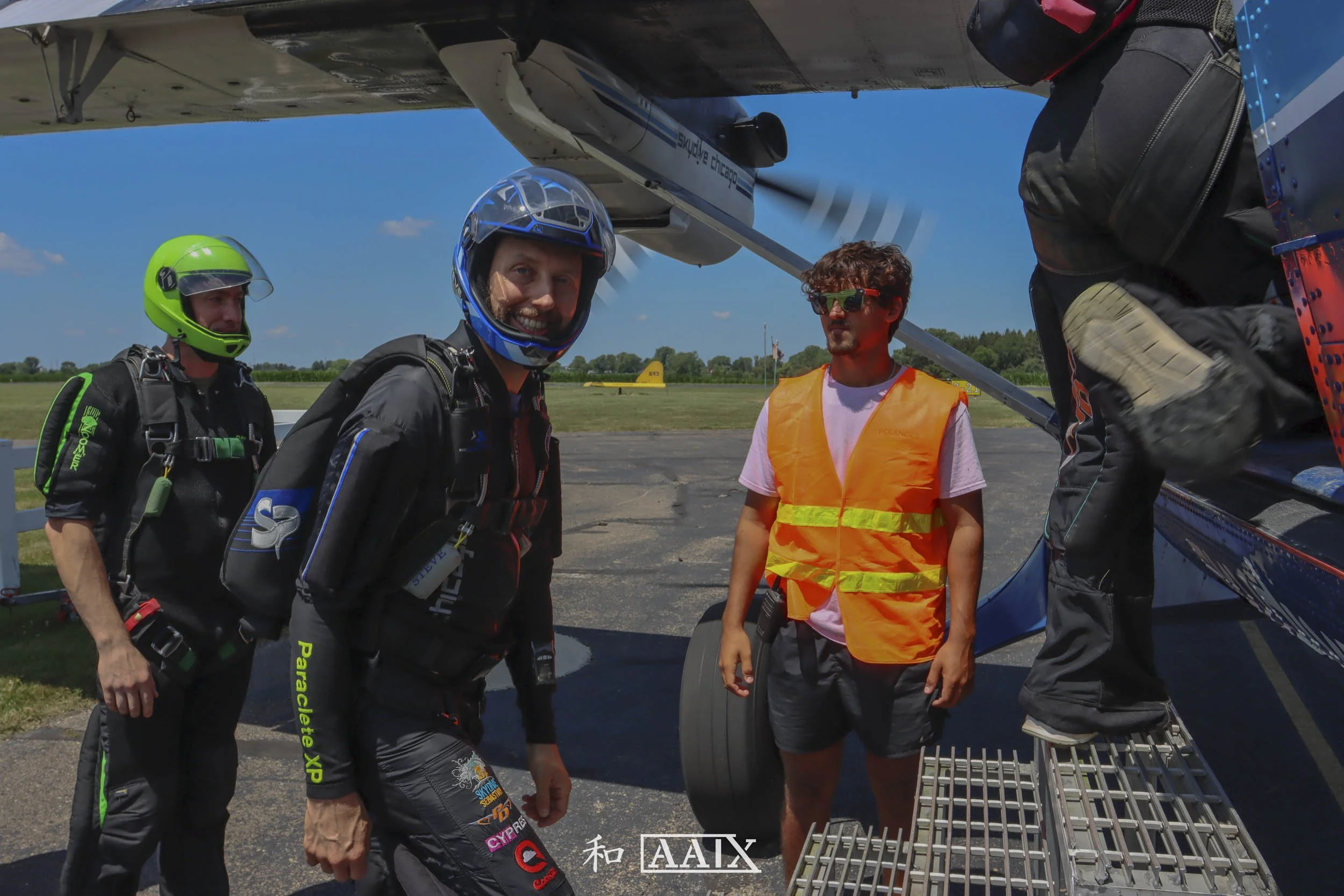 Three skydivers dressed in jumpsuits and helmets standing near an airplane on a grassy airfield, preparing for a jump under clear blue skies.
