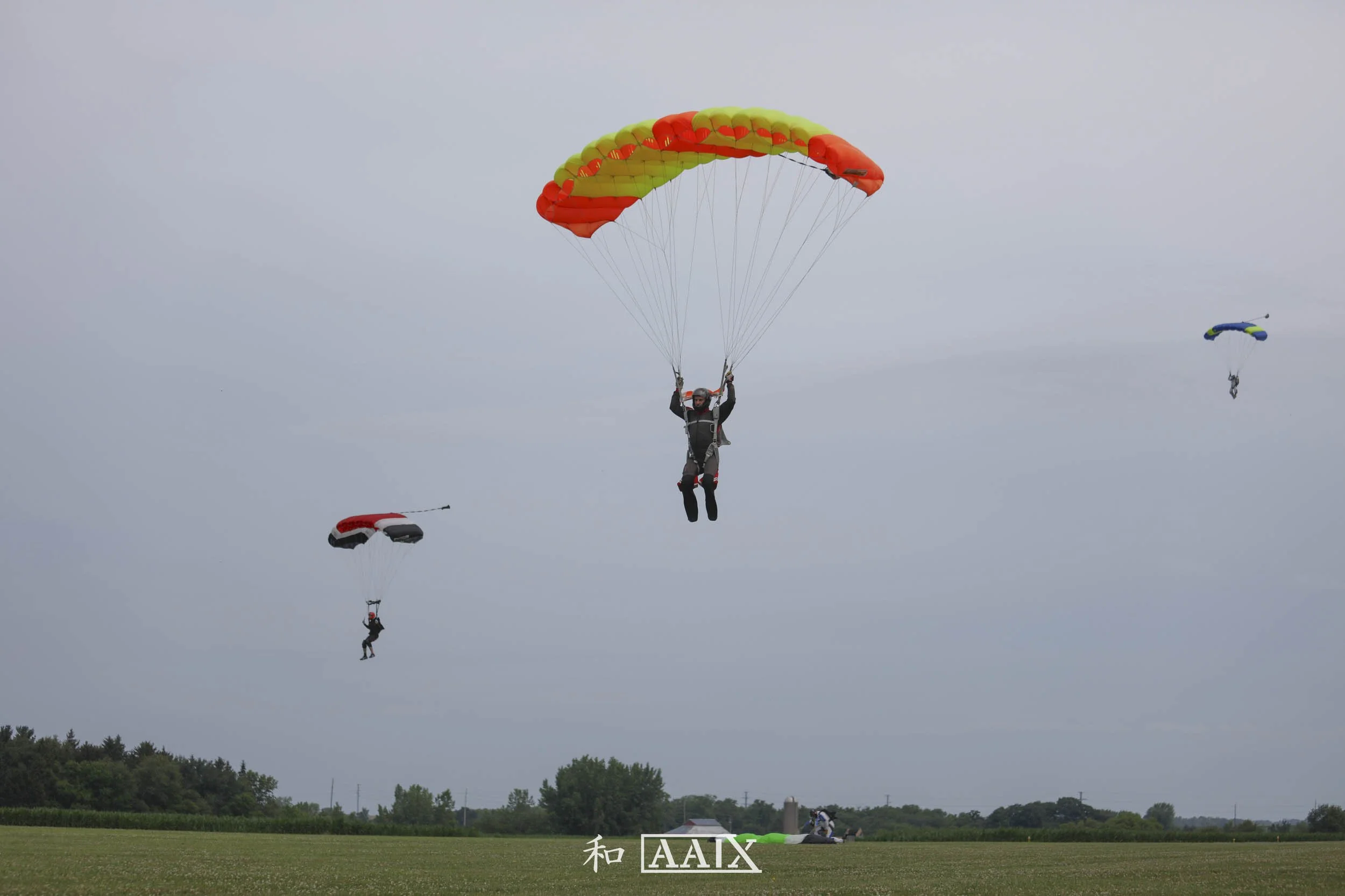 Three people paragliding over a grassy field against a cloudy sky, with trees in the background, and a person on the ground preparing a parachute.