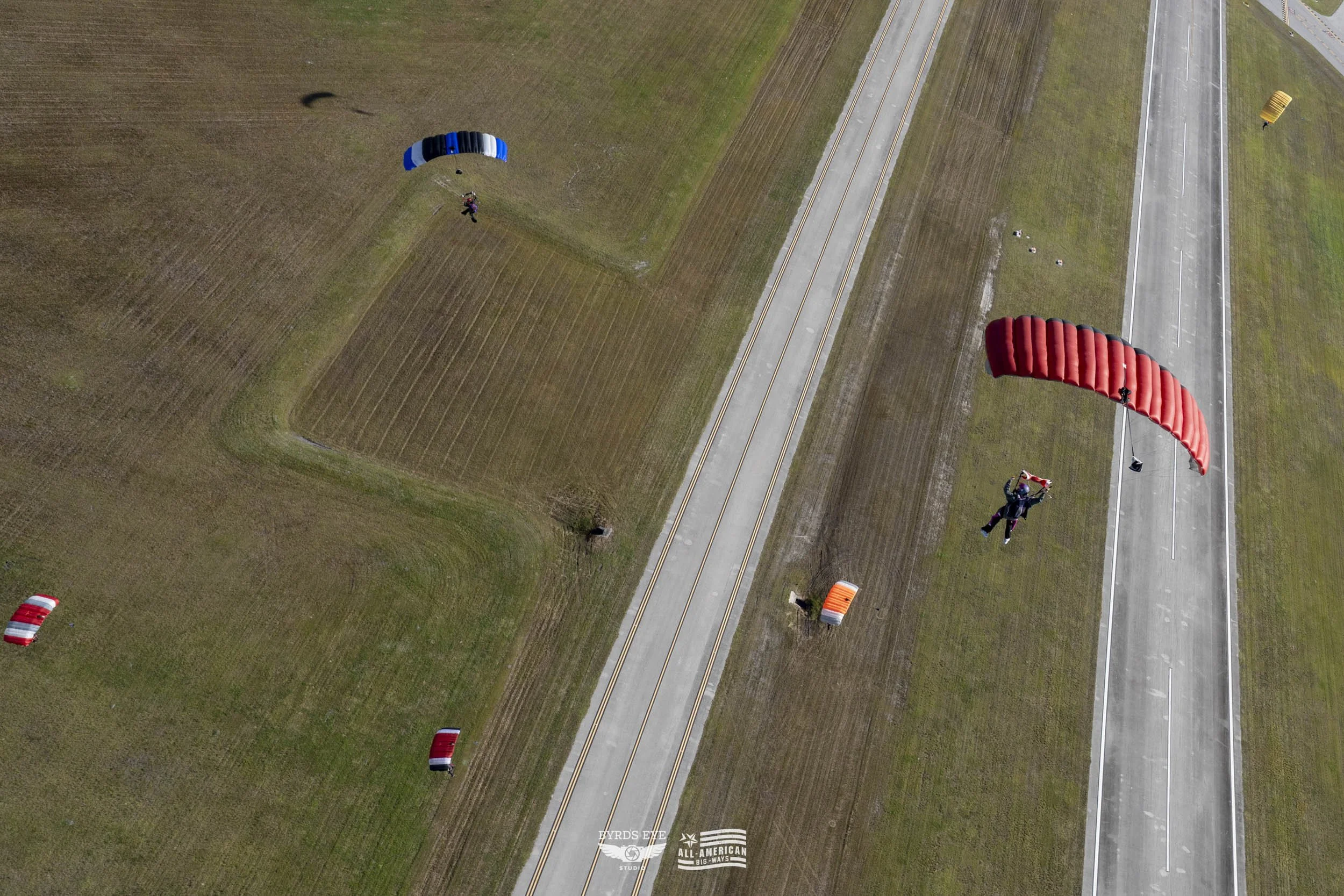 Two skydivers descending with parachutes over an open field near a highway, with smaller parachutes on the ground nearby.