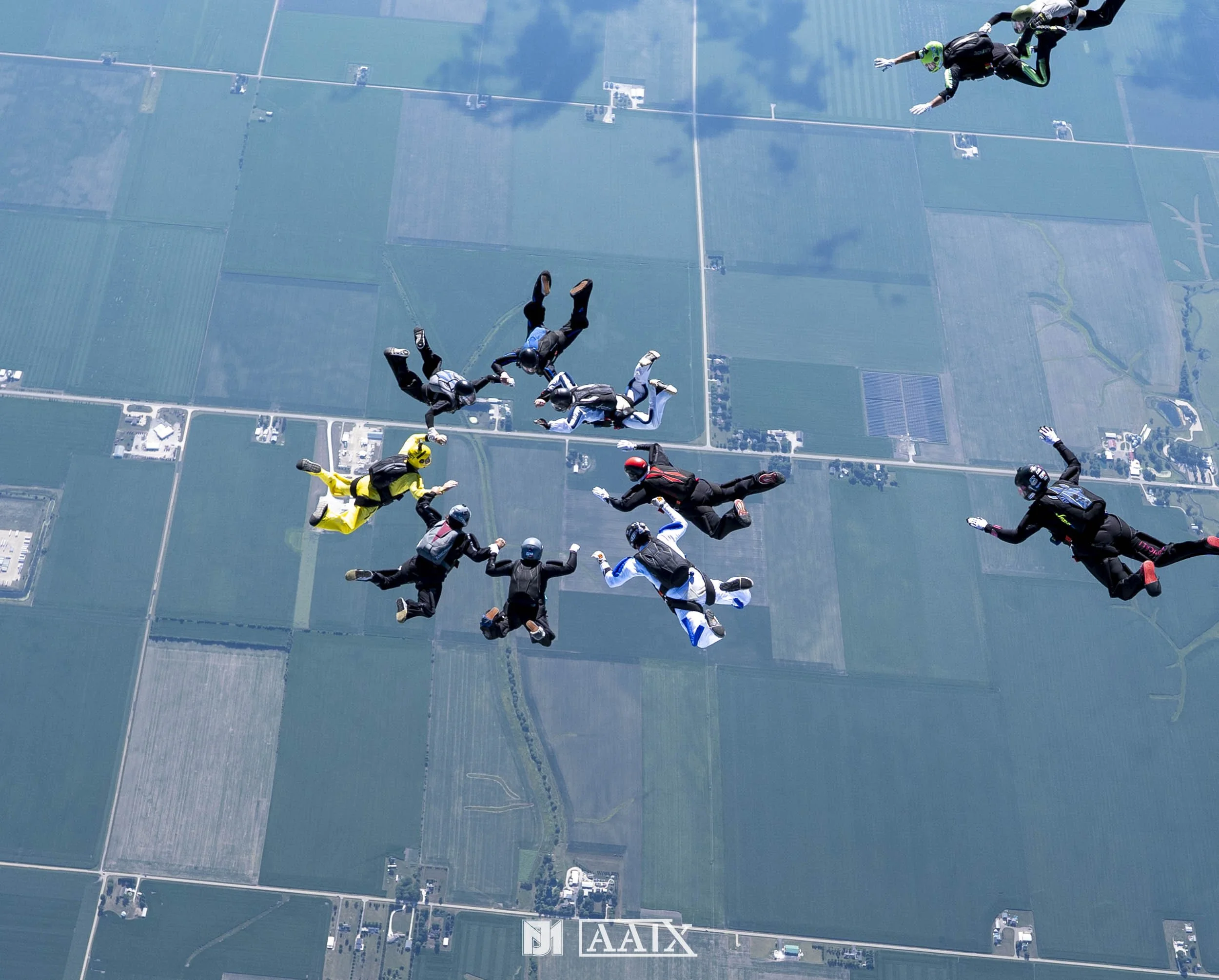 Group of skydivers in freefall over a rural landscape with fields and roads, many wearing jumpsuits and helmets.