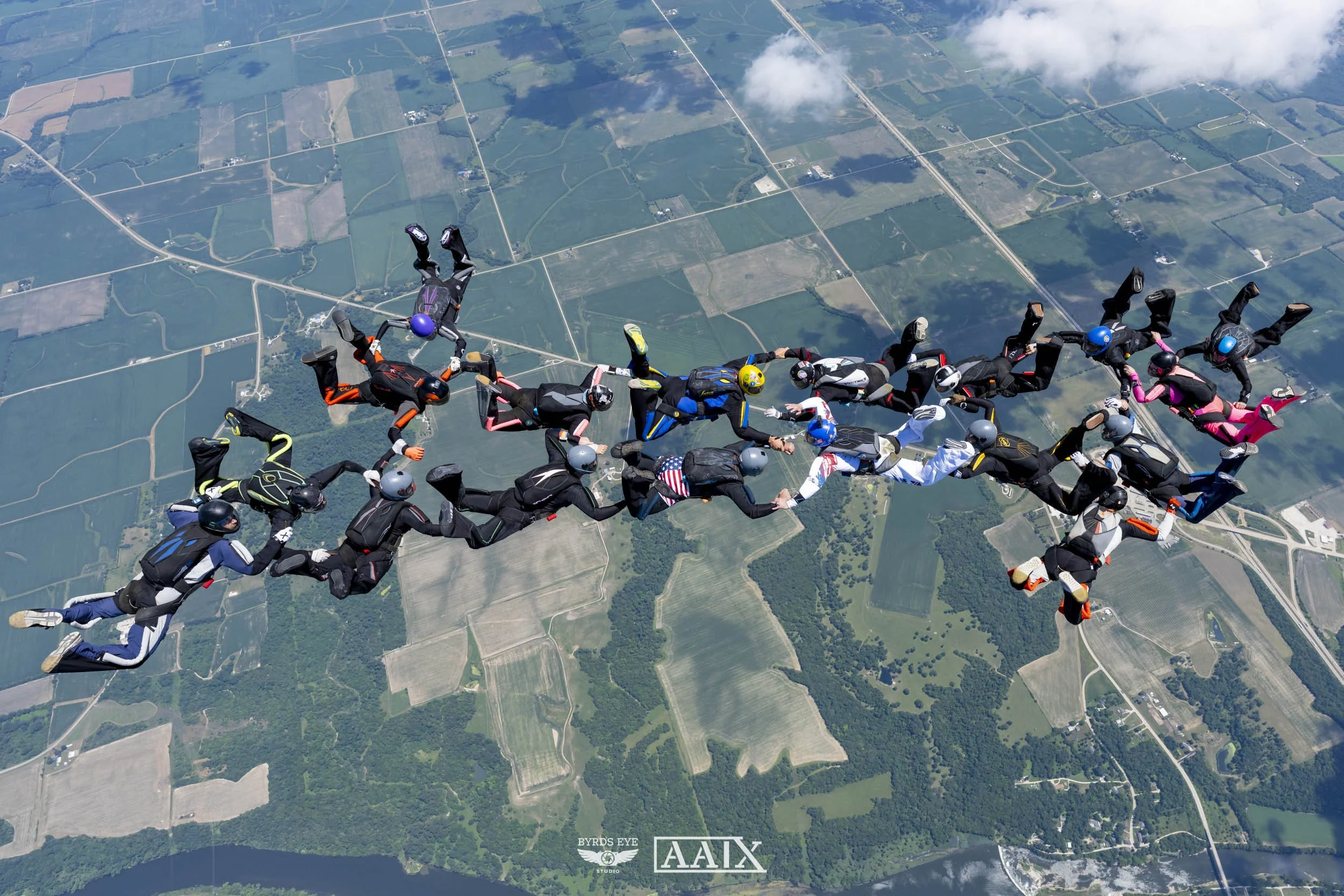 Group of skydivers in free fall over a patchwork of fields and rivers, holding hands in a line, with clouds overhead.