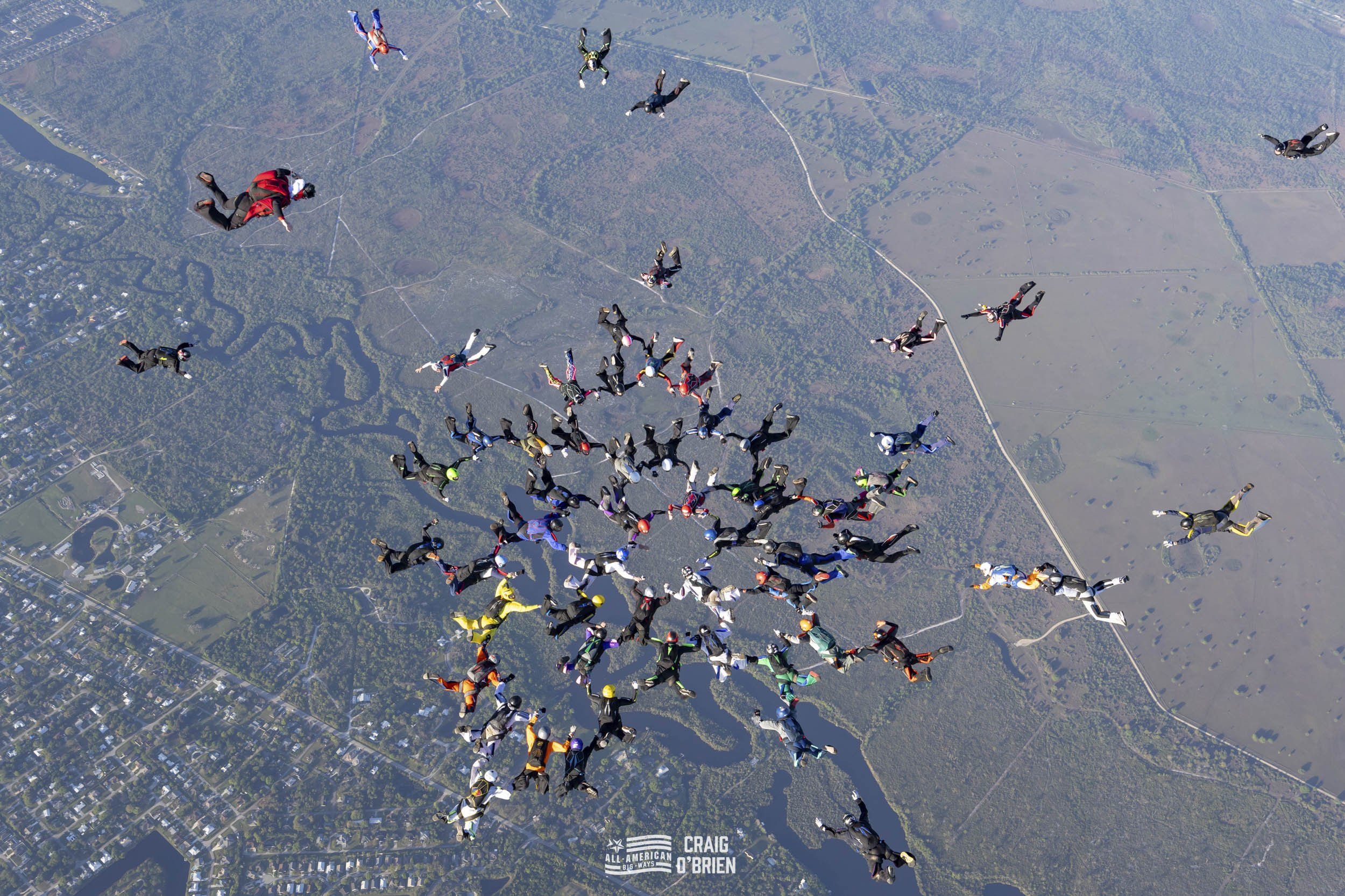 A group of skydivers in free fall above a landscape with rivers, trees, and fields, forming a circular formation.