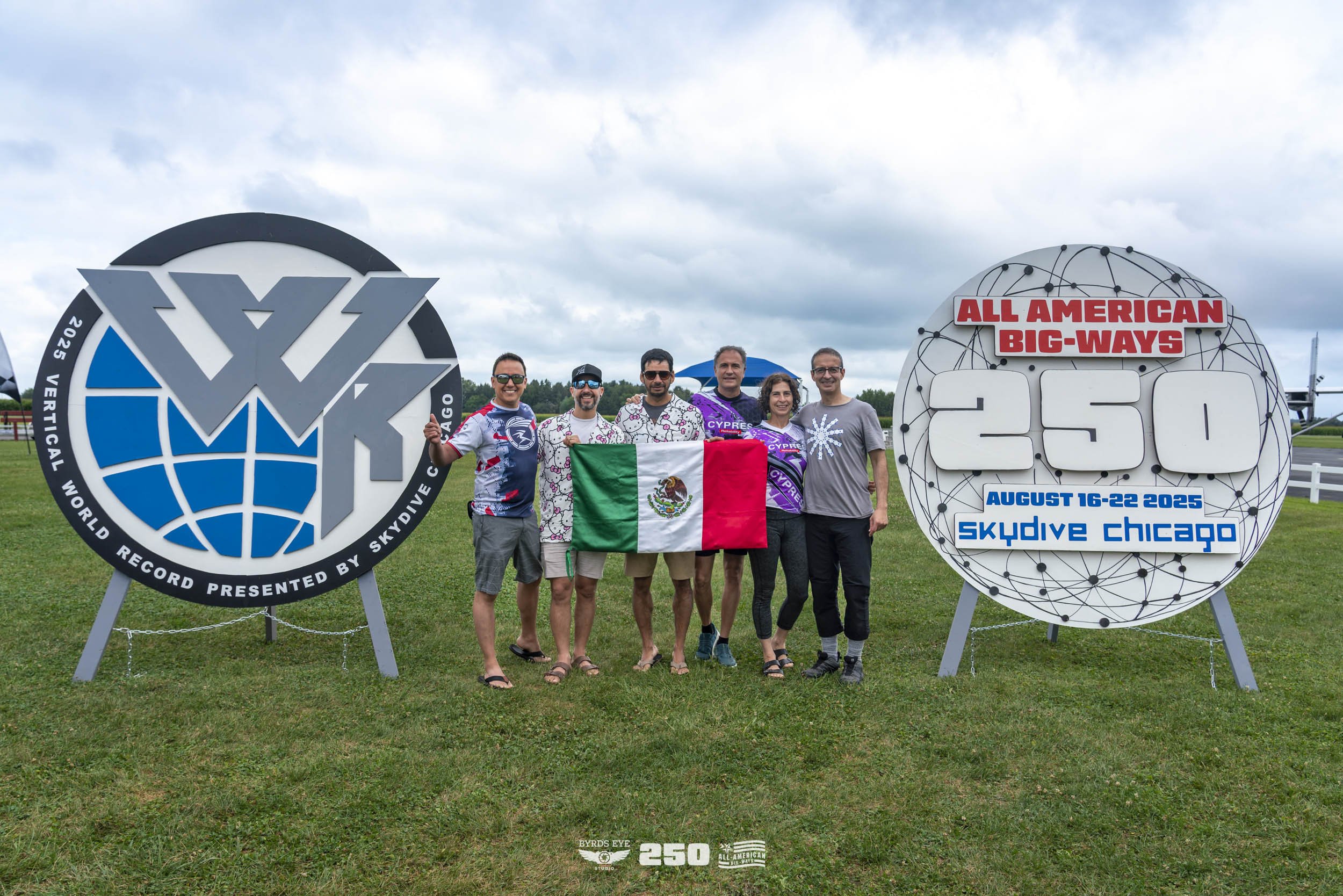 Group of six people standing outdoors on grass, holding a Mexican flag, beside large signs indicating a world record attempt for skydiving, with a cloudy sky overhead.