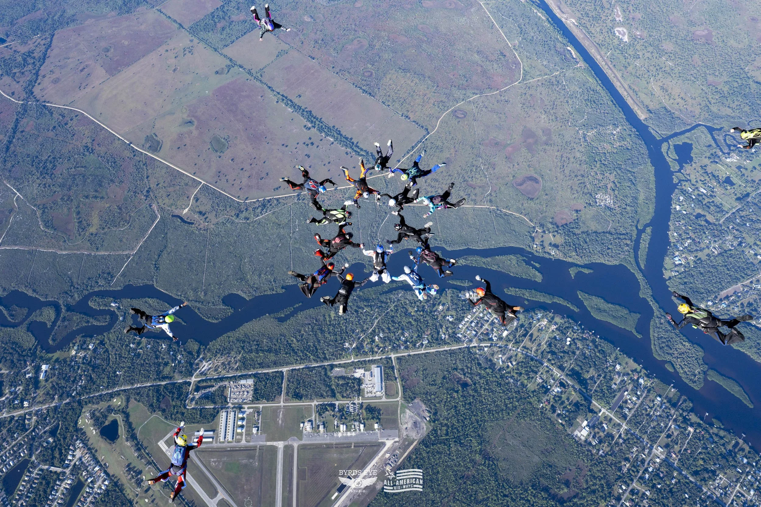 Skydivers free-falling in a group over a landscape with rivers, forests, and a small town.