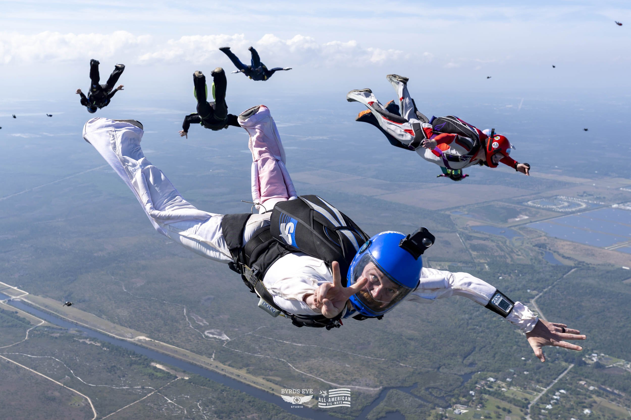 Group of five skydivers free-falling in the sky over a landscape with water, fields, and roads, with one skydiver in the foreground making a peace sign.