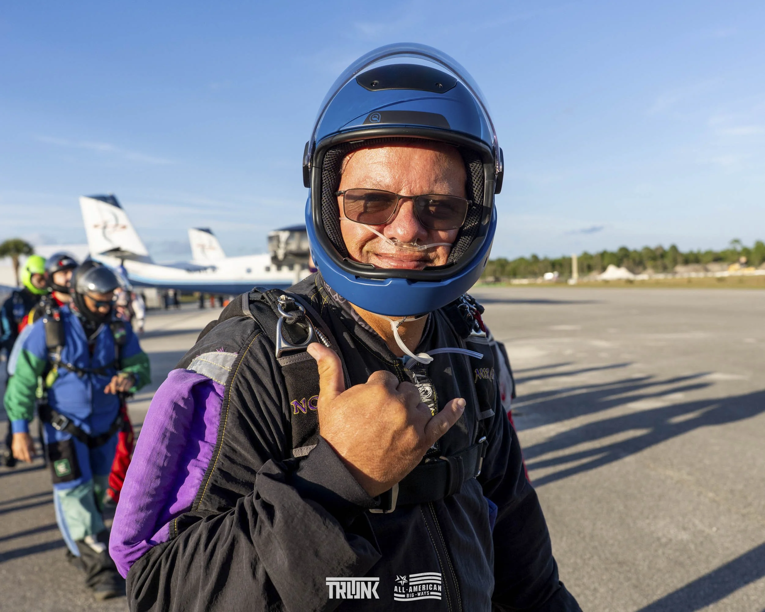 Skydiver wearing a blue helmet and sunglasses giving a rock on hand gesture, with other skydivers and airplanes in the background on an airport tarmac.