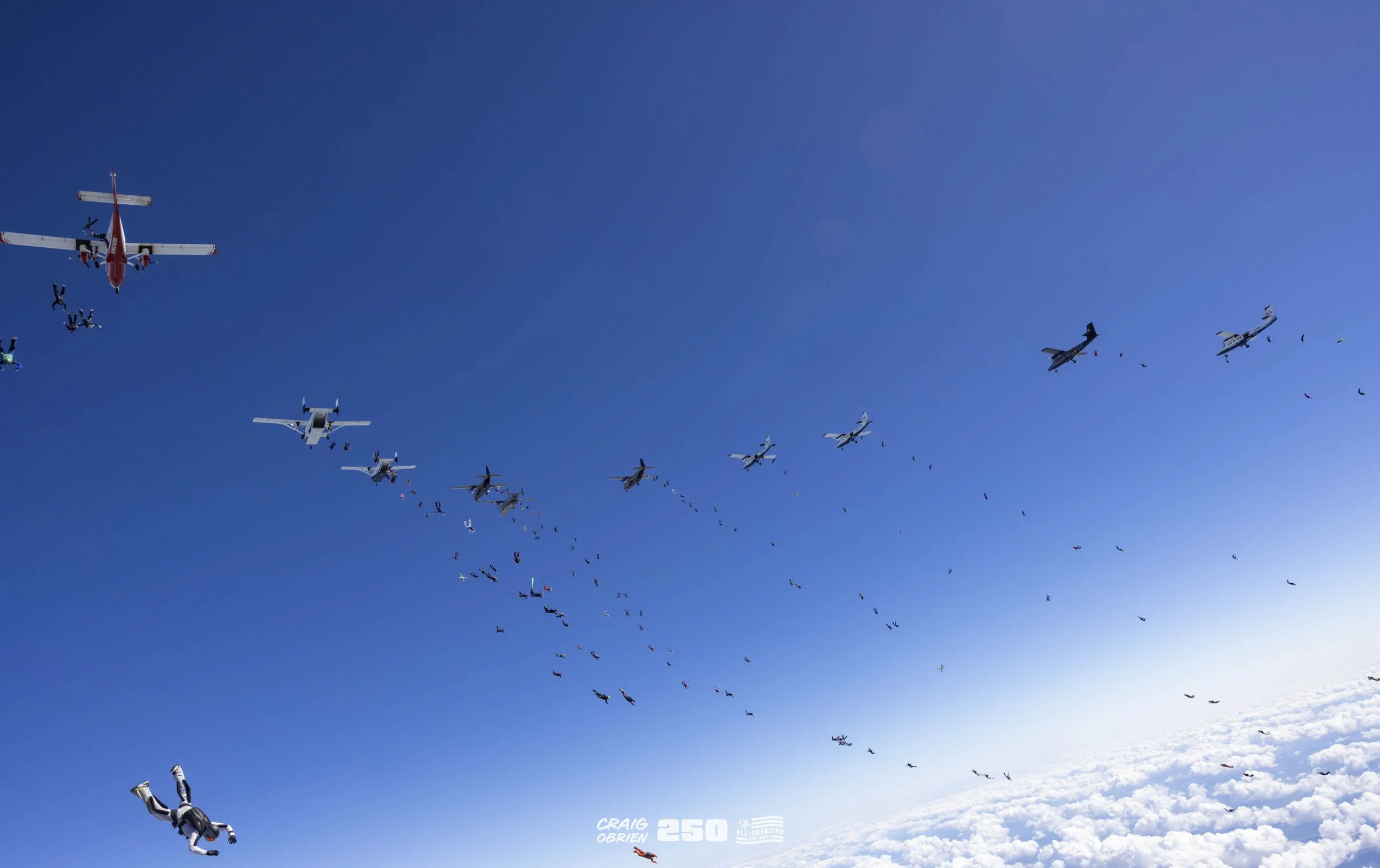 Skydivers free-falling from multiple airplanes in a blue sky above clouds.