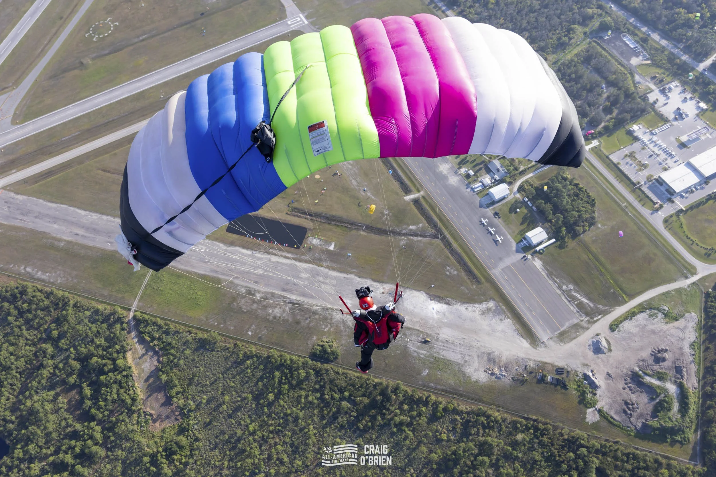 Person paragliding with a multi-colored wing over a landscape of roads, buildings, and forest.