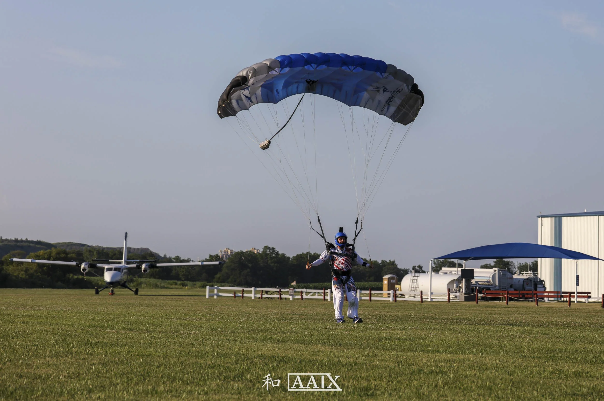 Person with a helmet and jumpsuit landing with a parachute in an open grassy field near an airplane and airfield structures.