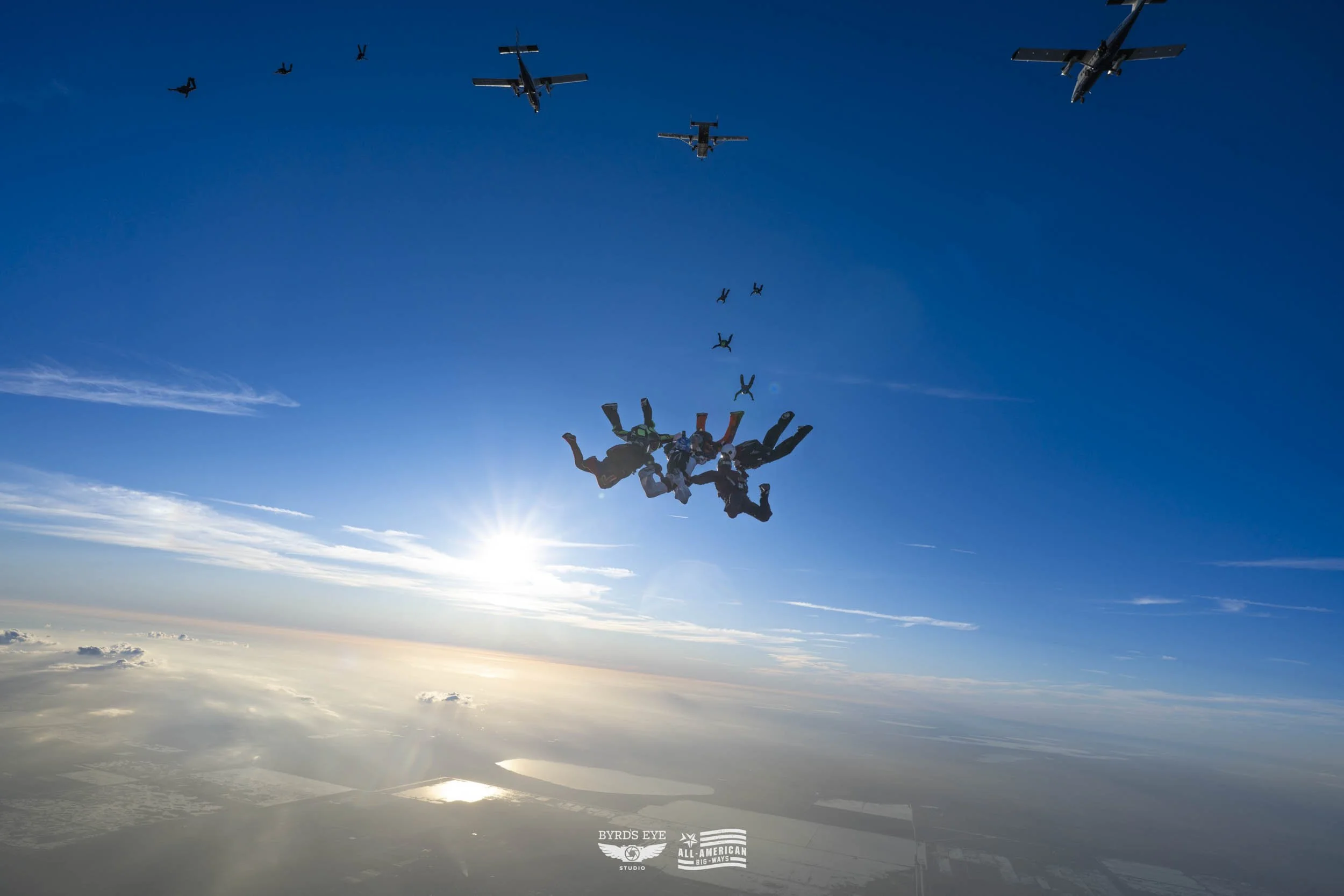Skydivers mid-free fall formation with airplanes above, set against a blue sky with the sun and clouds below.