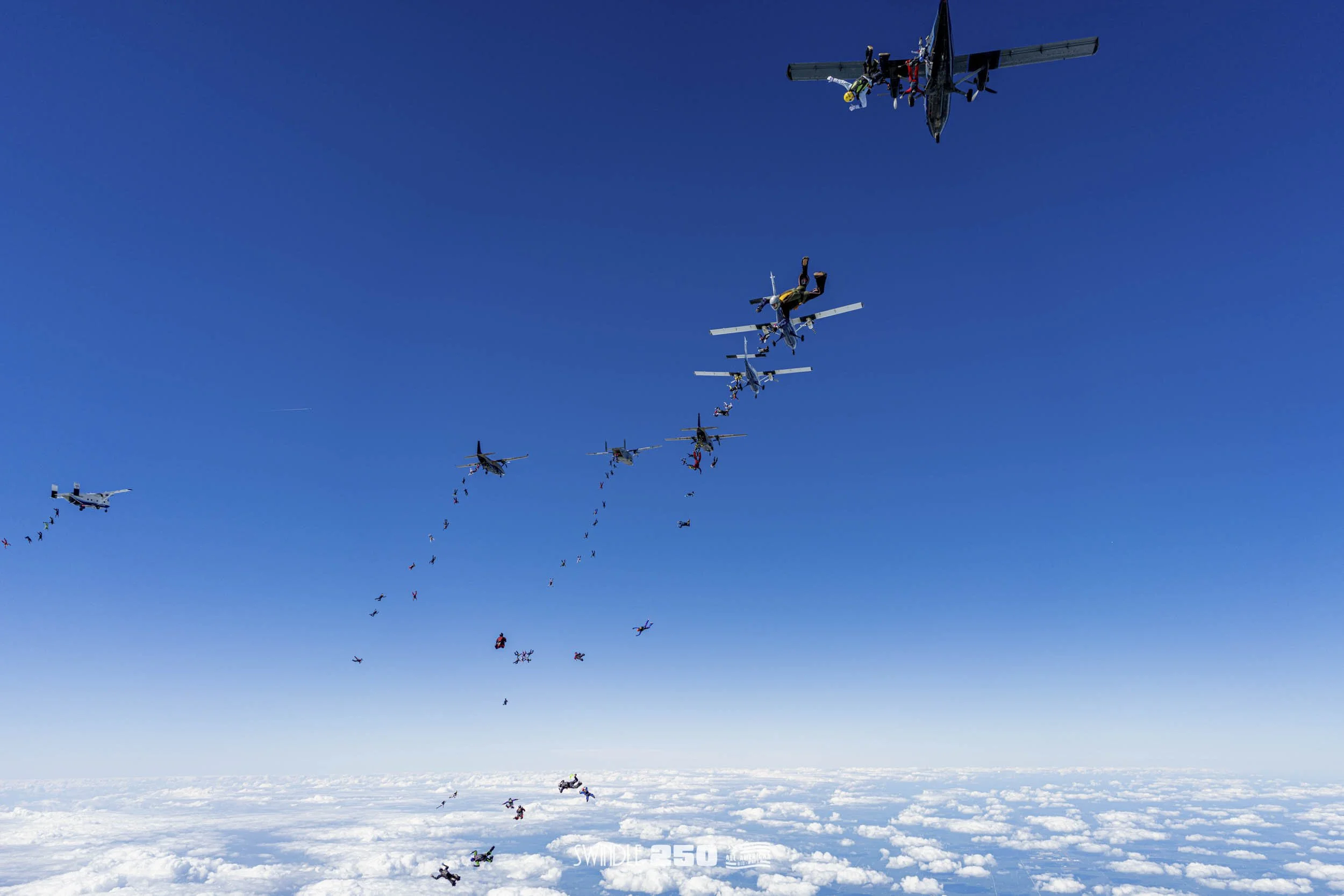 Skydivers jumping from a plane above the clouds with a clear blue sky in the background.
