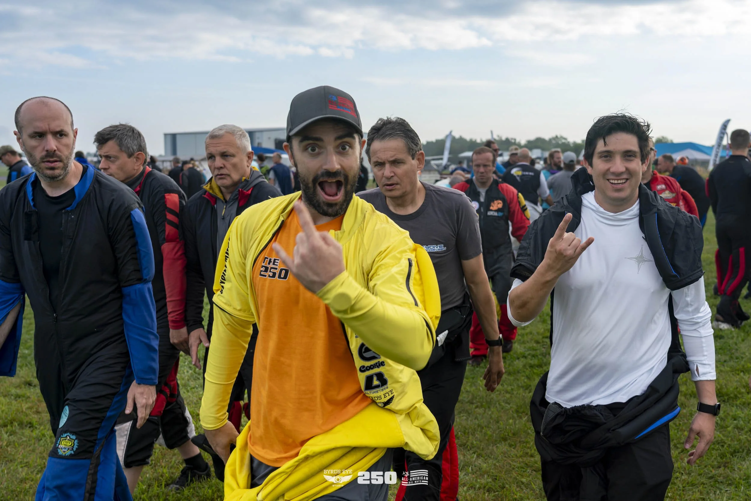 Group of men, some smiling and some surprised, walking outdoors on a grassy field during the day, with an airport or airfield in the background.