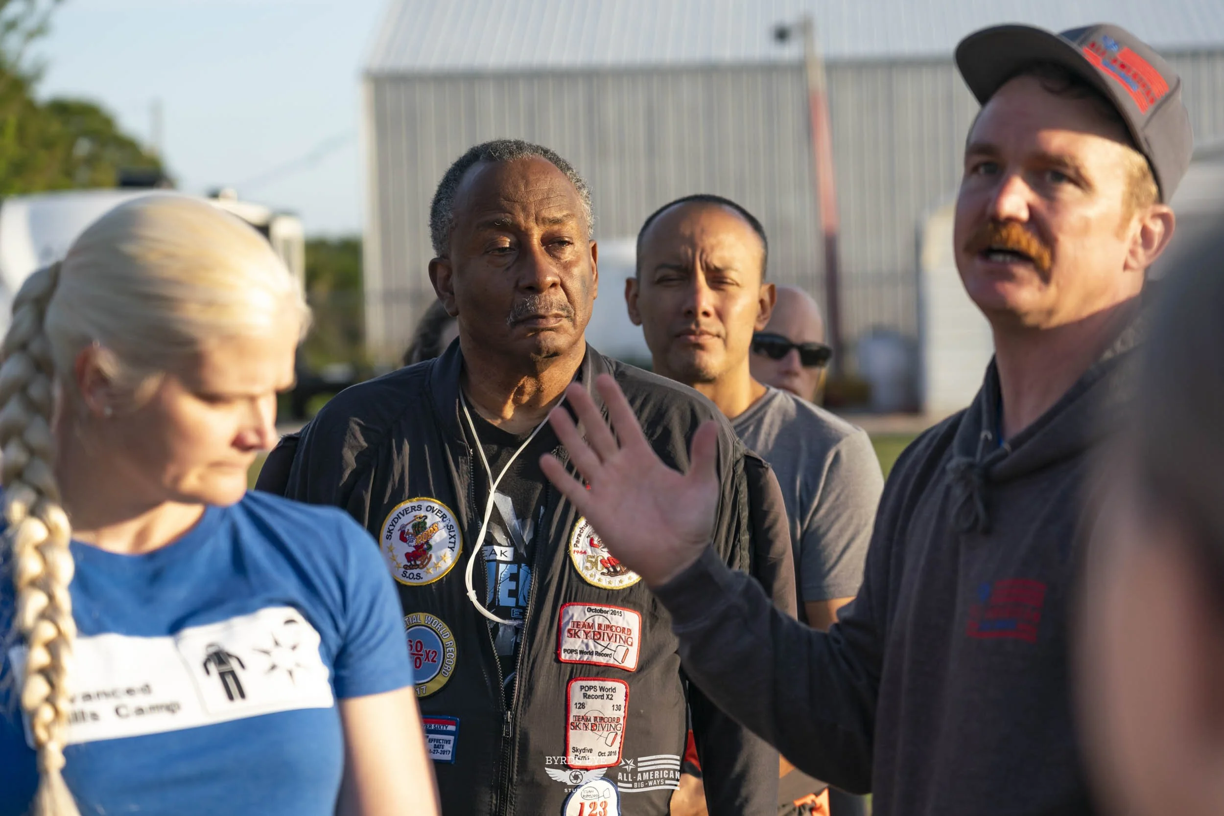 Group of people outdoors, with one man gesturing with his hand, others listening, and several wearing badges and casual clothing.